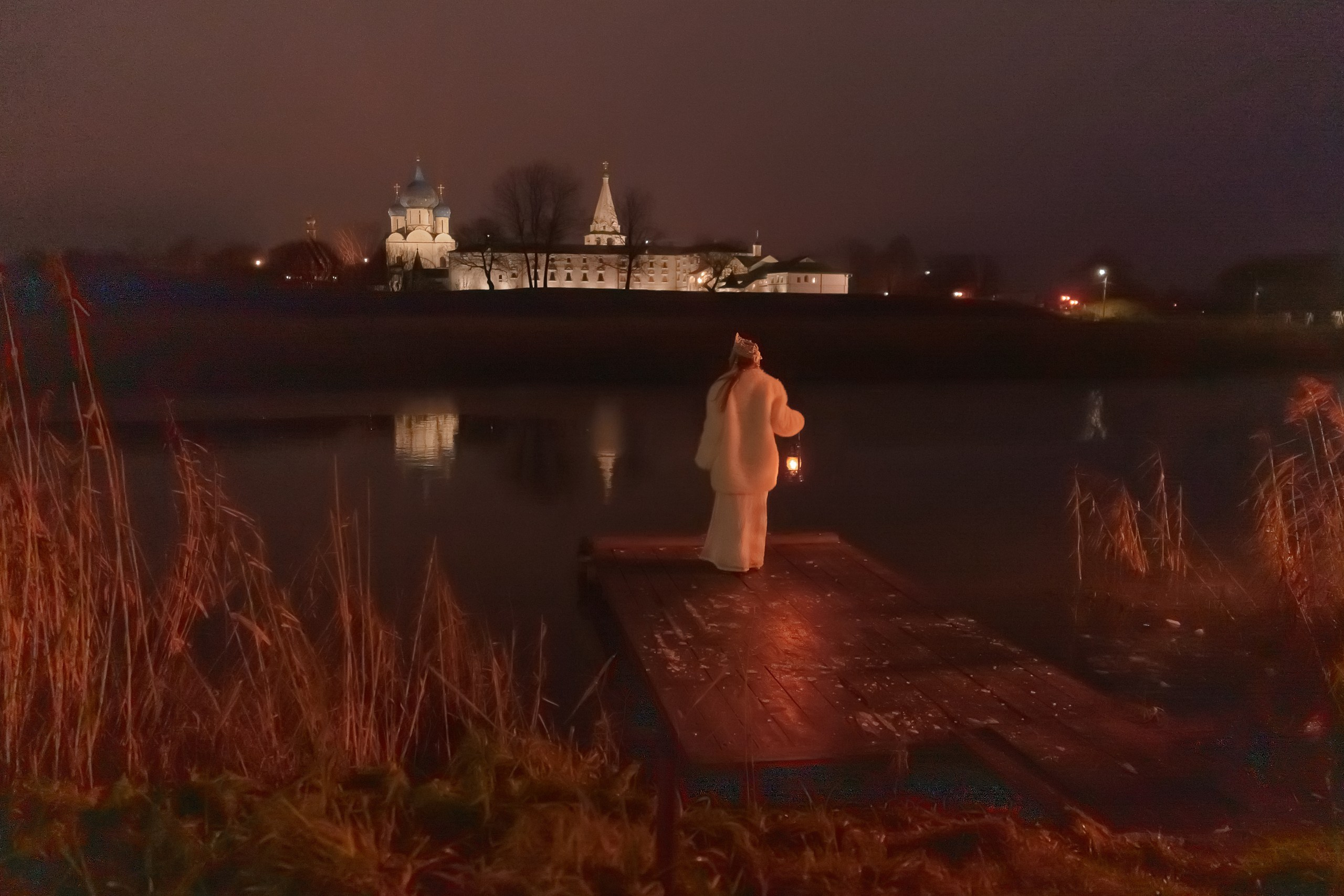 A girl in a white Russian folk costume with a kerosene lamp in the evening against the background of a temple by the river