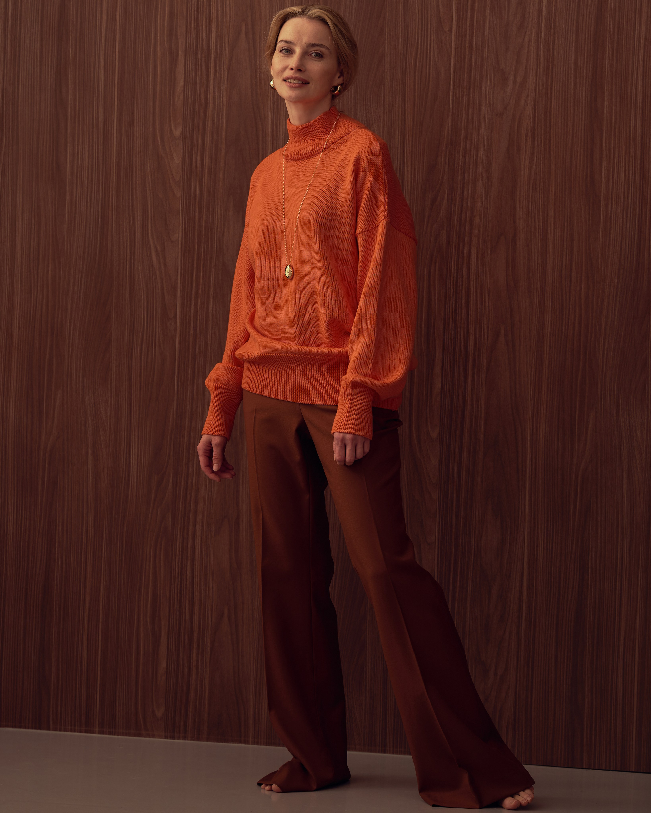 Съемка лукбука, девушка в оранжевой ower size свитере и коричневых брюках стоит на фоне деревянной стены