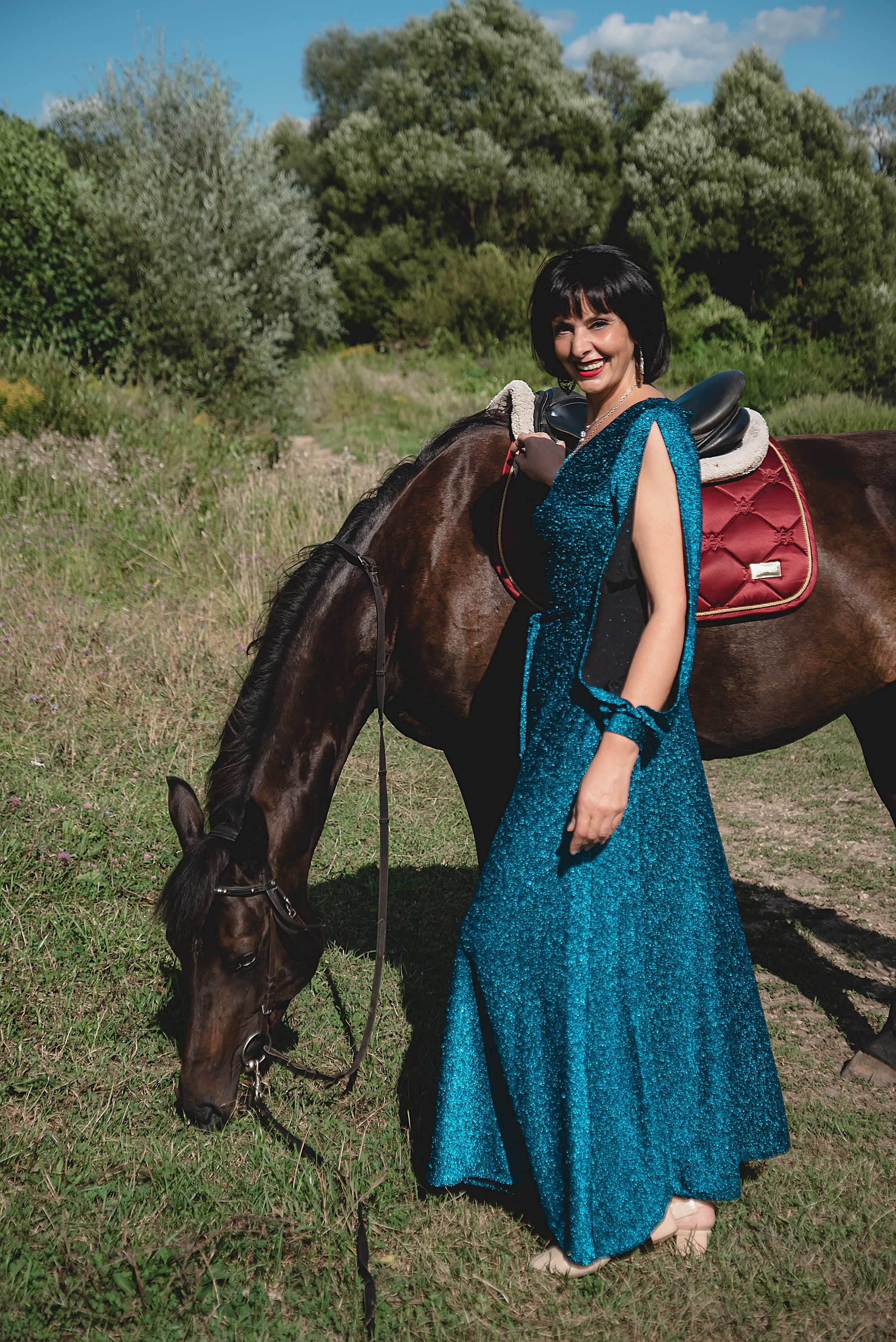 На фото женщина в зеленом платье стоит с лошадью на природе.