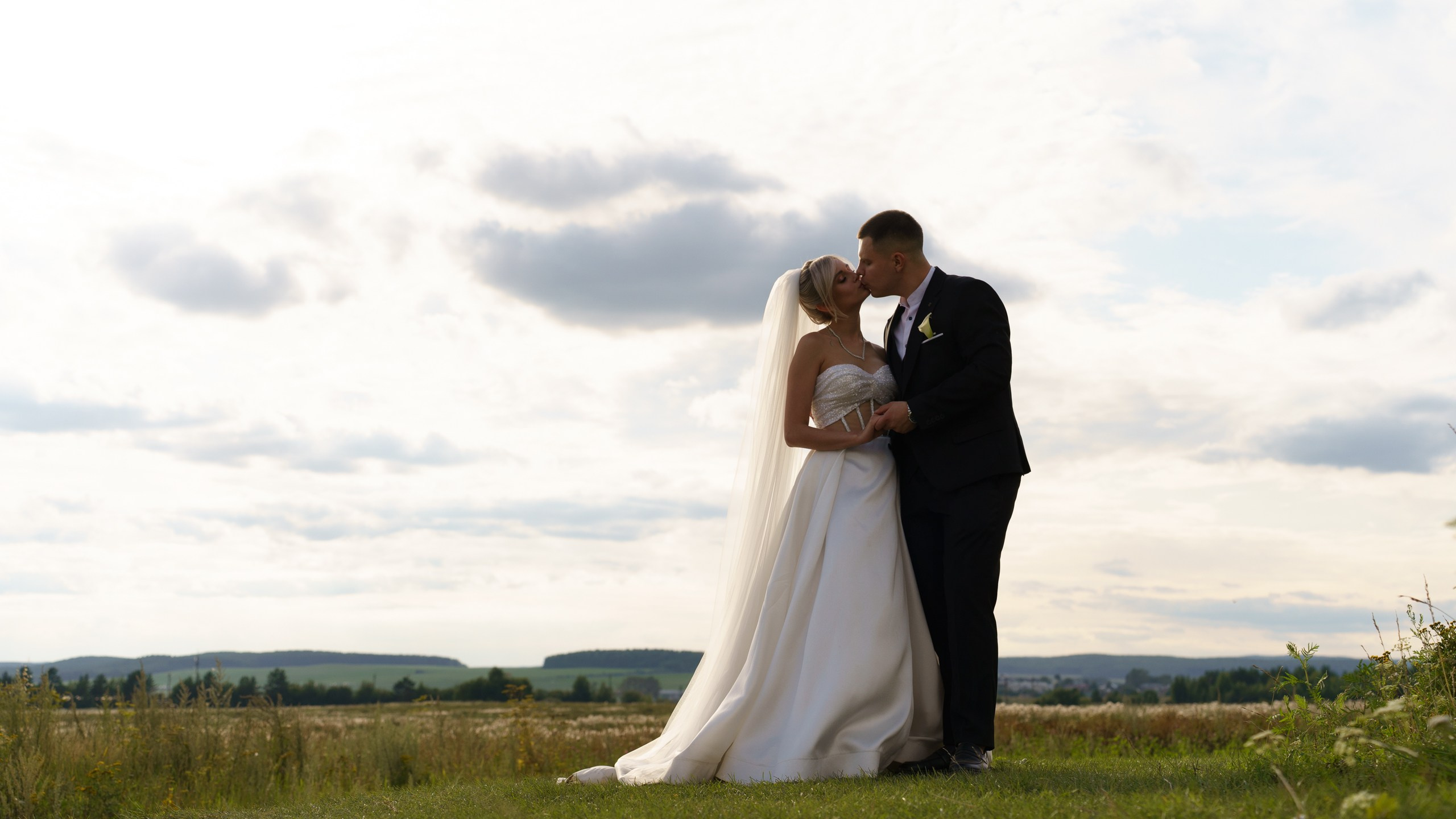 Свадебная фотосессия на природе: пара стоит на траве, жених нежно целует невесту. Вокруг — живописные холмы и бескрайнее небо | оригинальные свадебные фотографии Екатеринбург