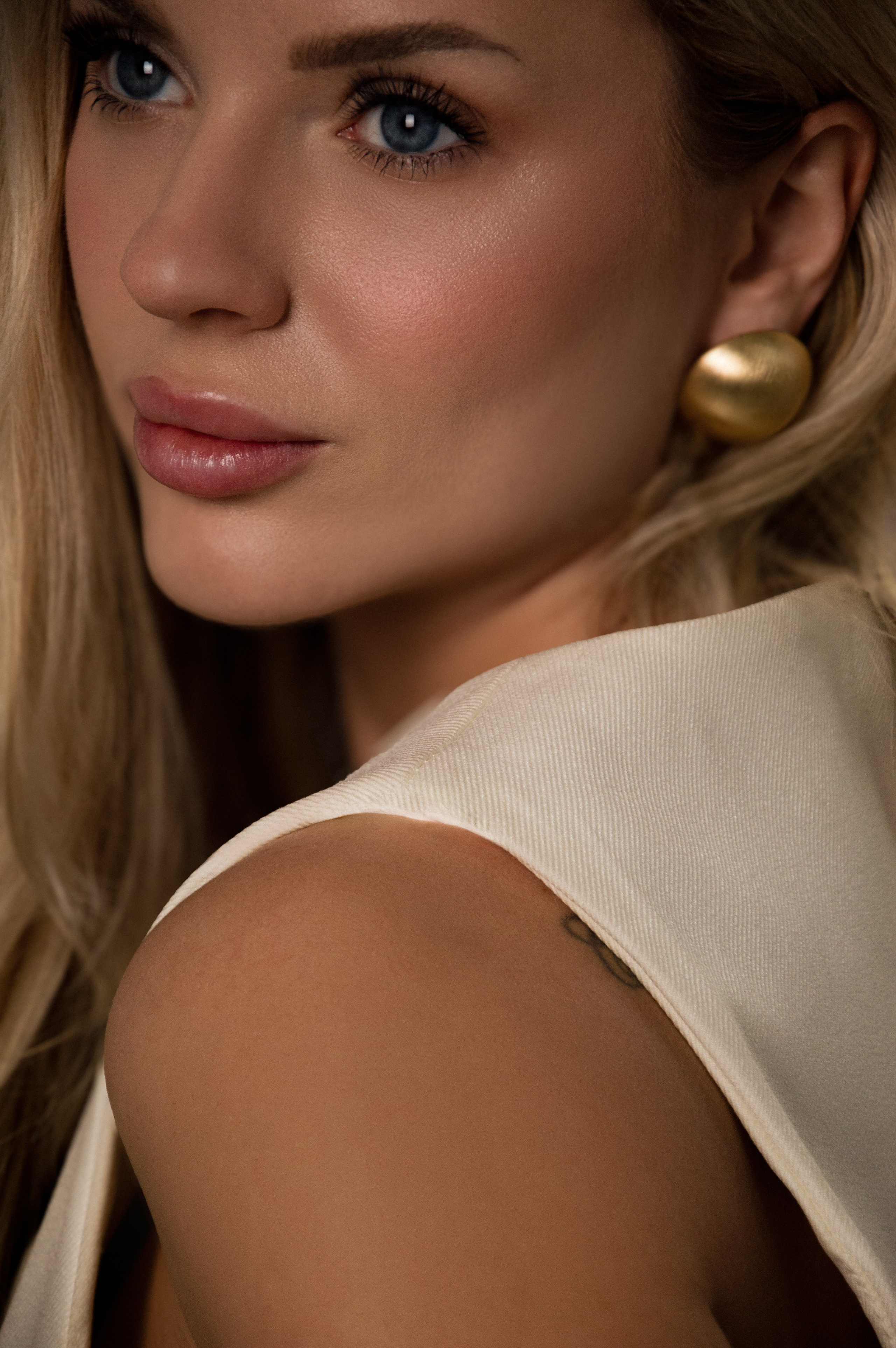 Игра света и текстуры: крупный план лица блондинки с выразительными глазами, дополненный крупными золотыми серьгами.