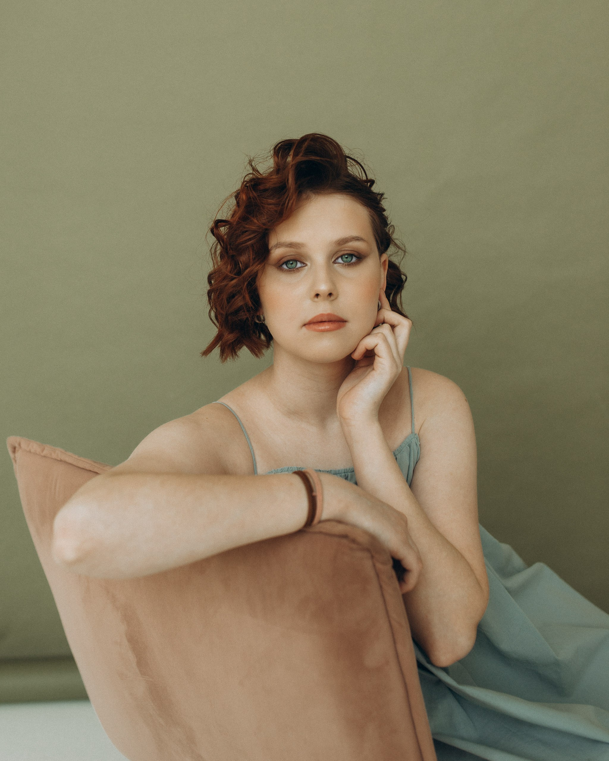 Портрет девушки с рукой у подбородка. Задумчивый взгляд, естественный макияж и голубое платье на оливковом фоне студии. Подростковая фотосессия
