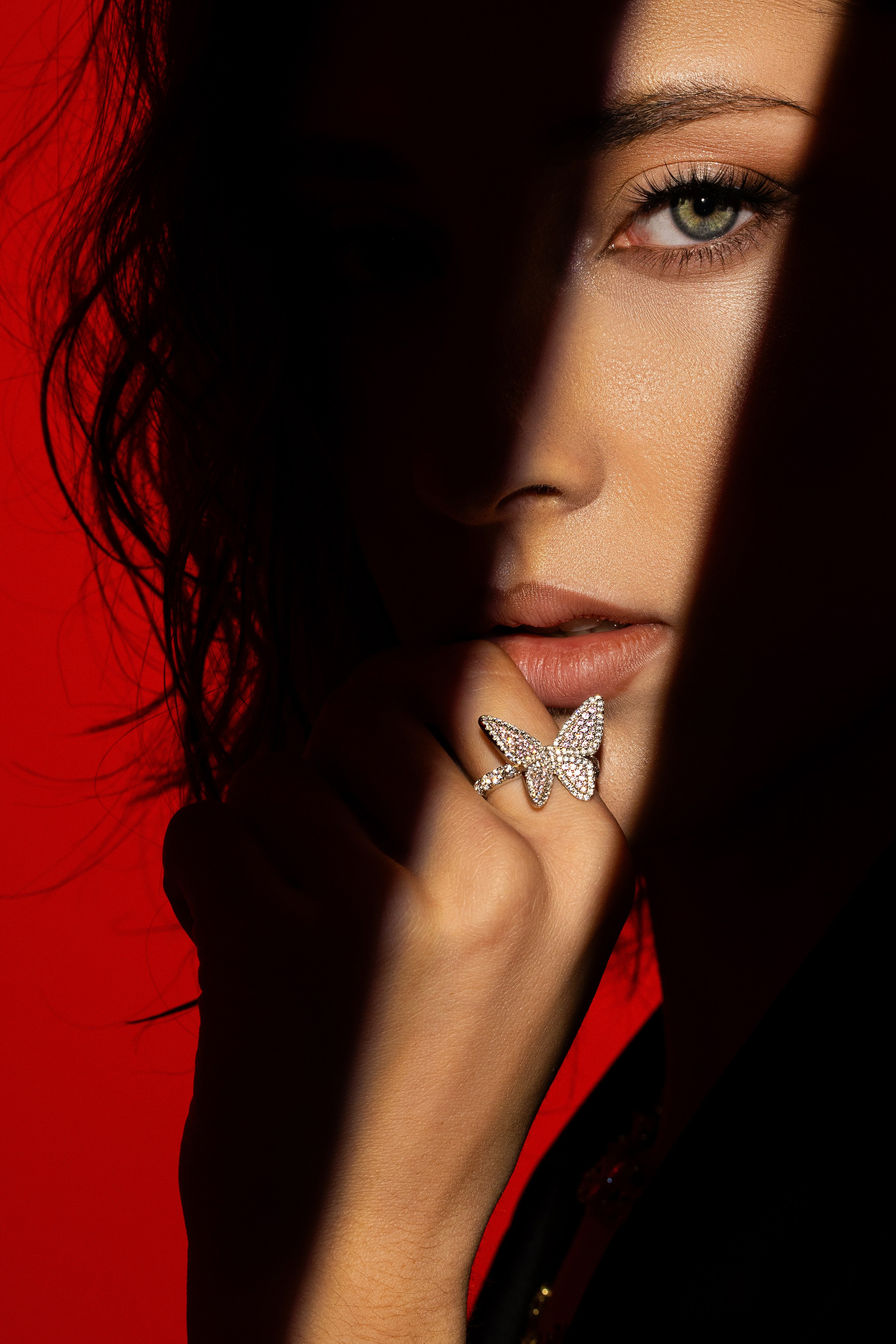 Съёмка для ювелирного бренда Aporro: крупный план женщины, на пальце которой сверкает кольцо с дизайном в виде бабочки, покрытое драгоценными камнями. Сильный контраст света и тени подчеркивает детали украшения, а яркий красный фон создаёт атмосферу драматичности и элегантности