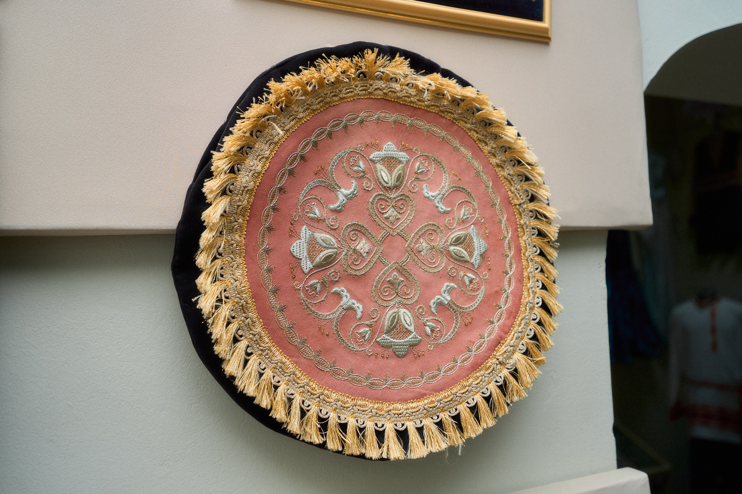 Круглое декоративное панно с традиционной торжокской золотошвейной вышивкой. Основной фон панно — розовый, с центральным узором из серебряных и золотых нитей, образующих сложный орнамент. Края панно украшены золотыми кистями, что придает изделию роскошный и торжественный вид