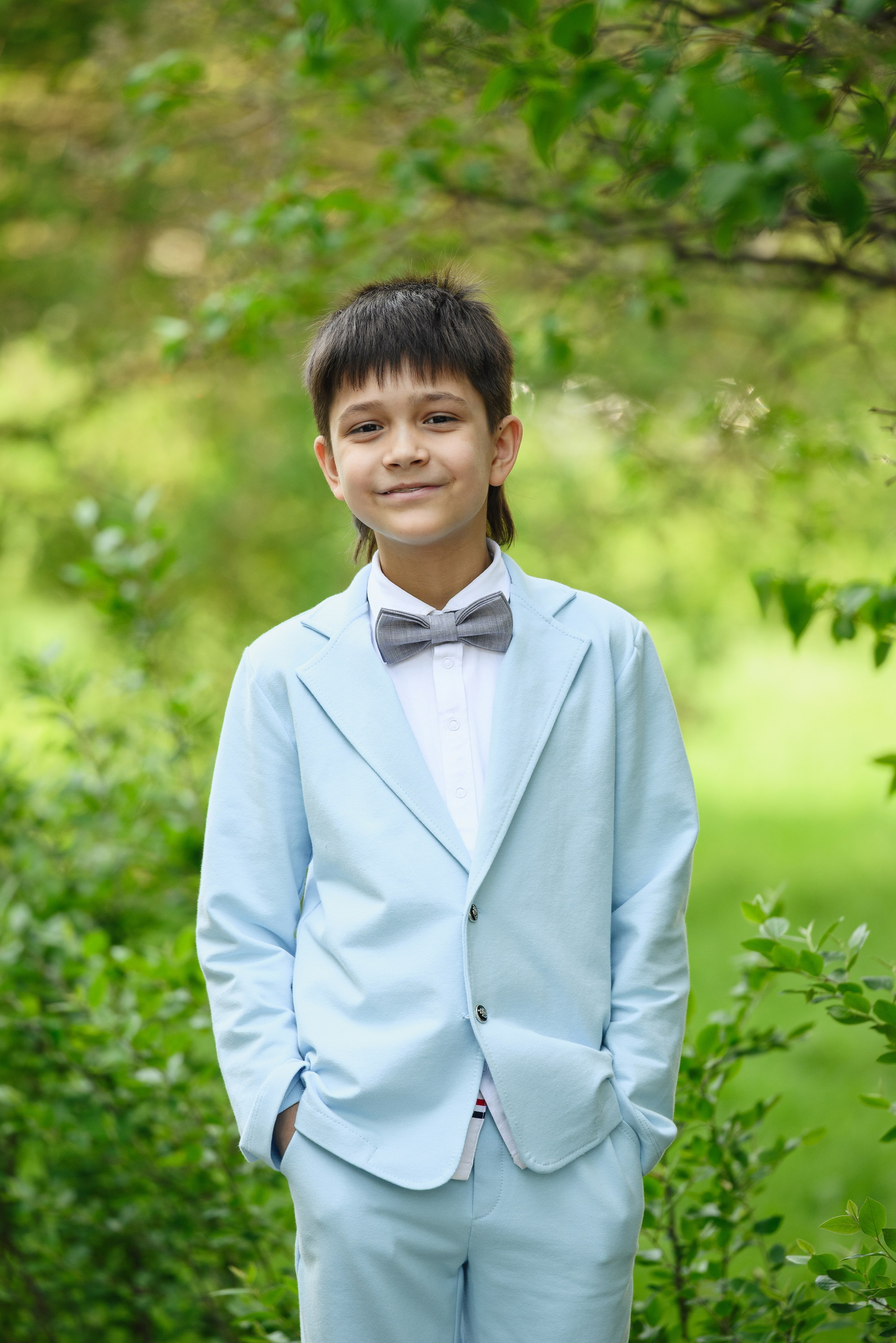 Мальчик в светлом костюме с бабочкой на фоне зелени