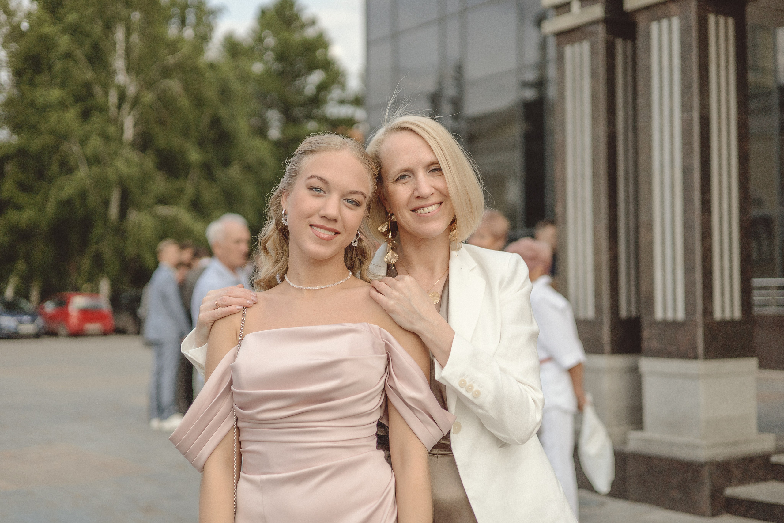 Мать и дочь тепло улыбаются у современного здания. На дочери розовое платье, а на матери белый блейзер. На заднем плане - деревья и люди.
