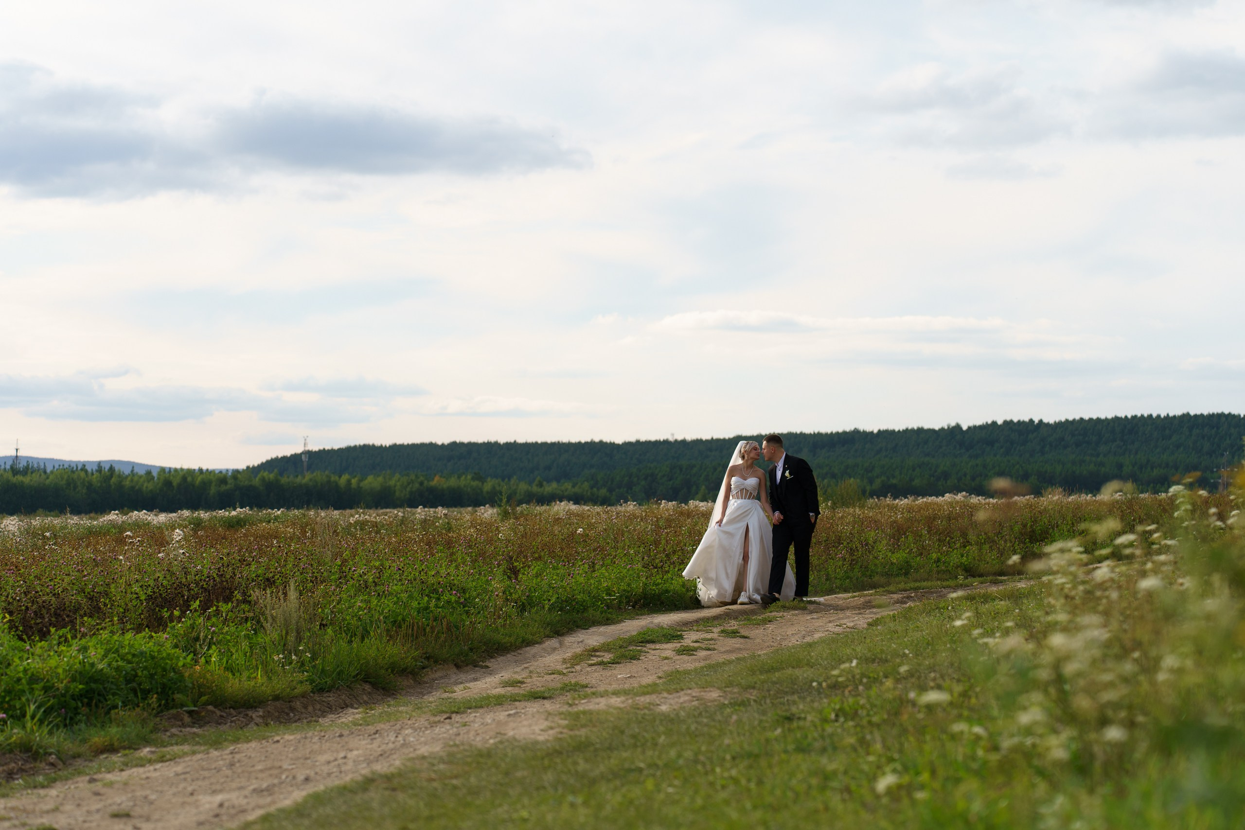 Свадебная пара на фоне природы: невеста в элегантном свадебном платье и жених в строгом костюме наслаждаются моментом, прогуливаясь по просёлочной дороге среди полевых цветов и зелени | свадебные фотографии на природе Екатеринбург