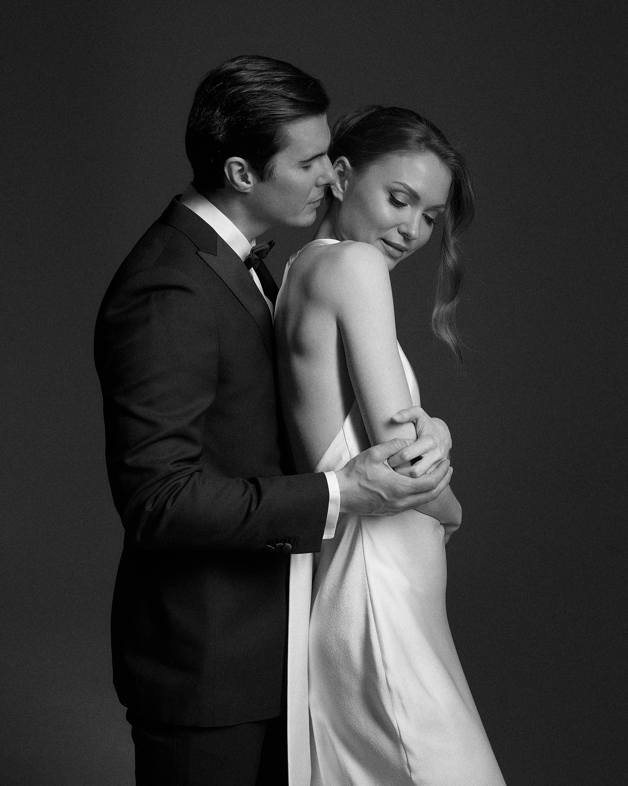 Он обнимает её так, будто хочет защитить от всего мира. Тёмный фон, минимум света — только на их лицах. Чувственный момент свадебной съемки, фотограф Дмитрий Цветков, СПб