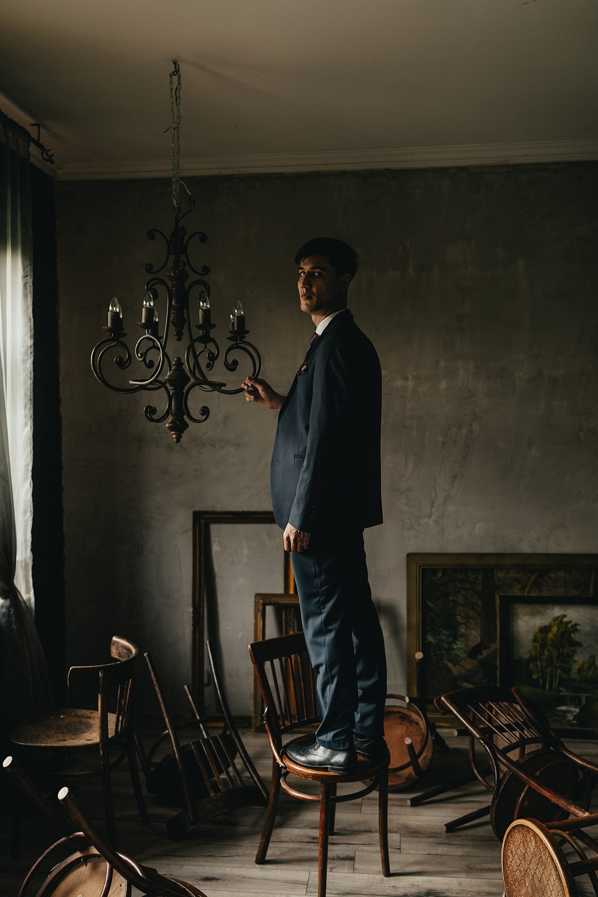 Мужчина в строгом костюме стоит на стуле, держа за люстру в старинной комнате с обстановкой из прошлого века