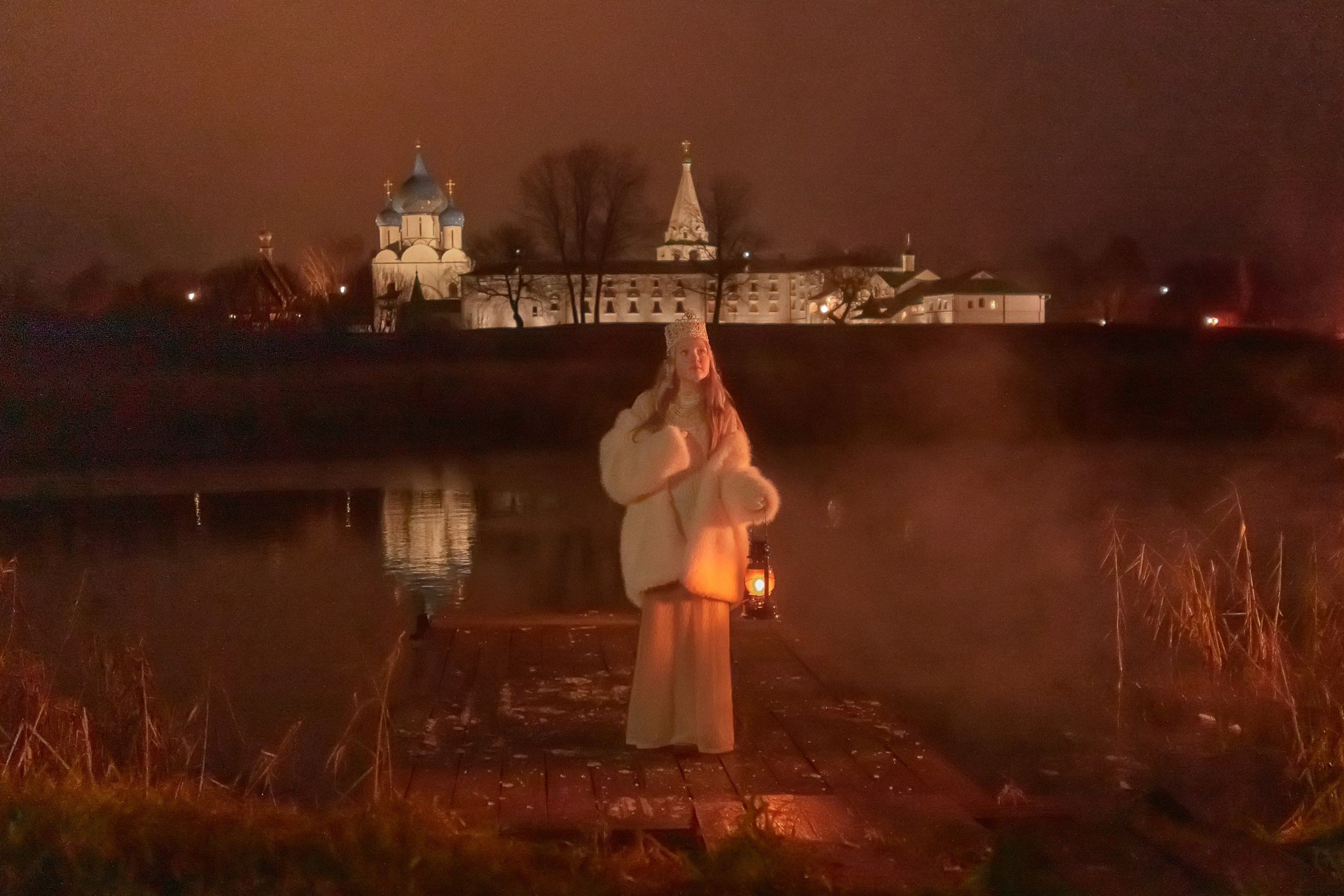 A girl in a white Russian folk costume with a kerosene lamp in the evening against the background of a temple by the river