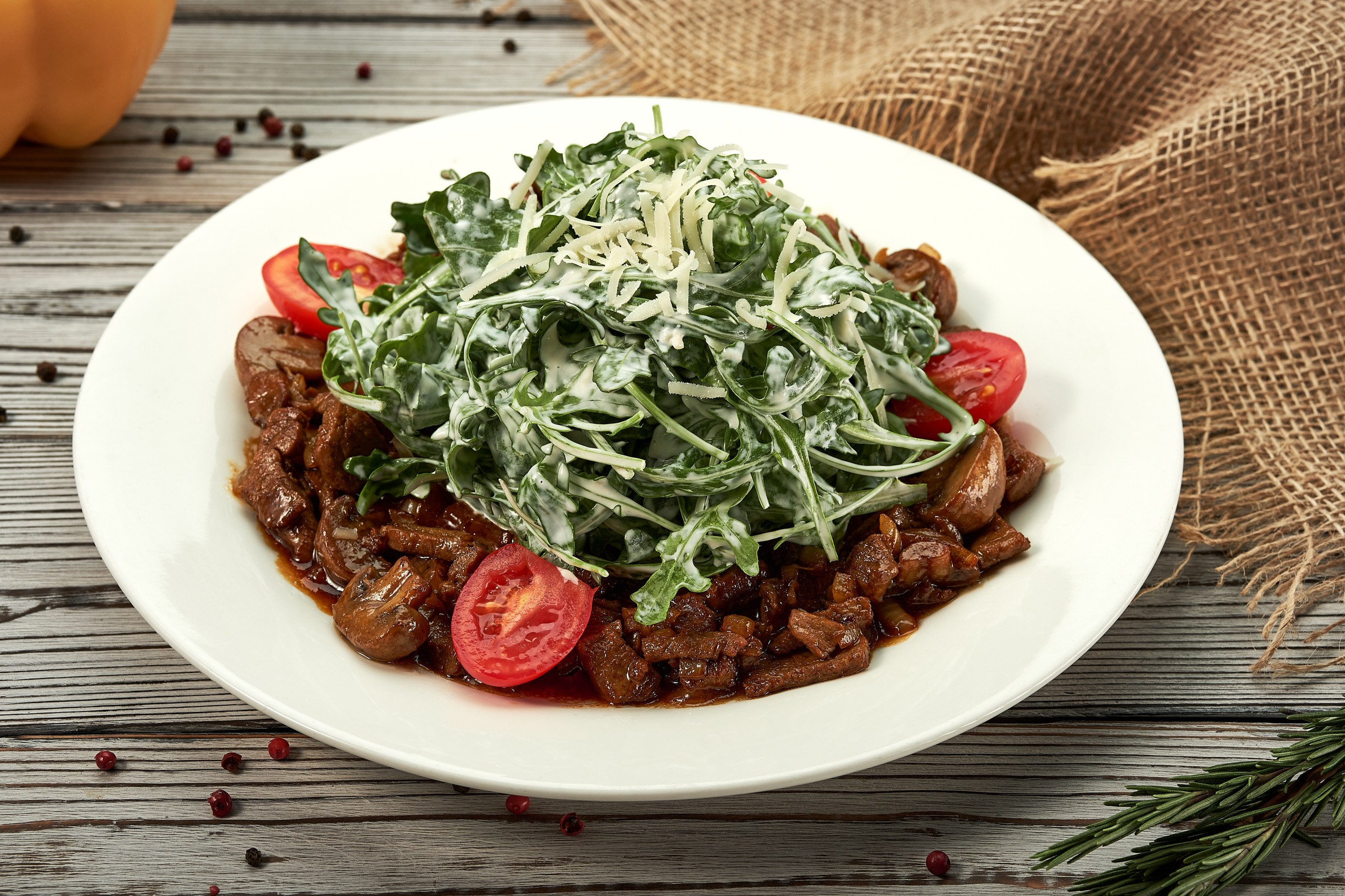 Горячая закуска с мясом и шампиньонами в соусе, украшенная свежей рукколой, тёртым сыром и томатами. Подача на белой тарелке на деревянной поверхности