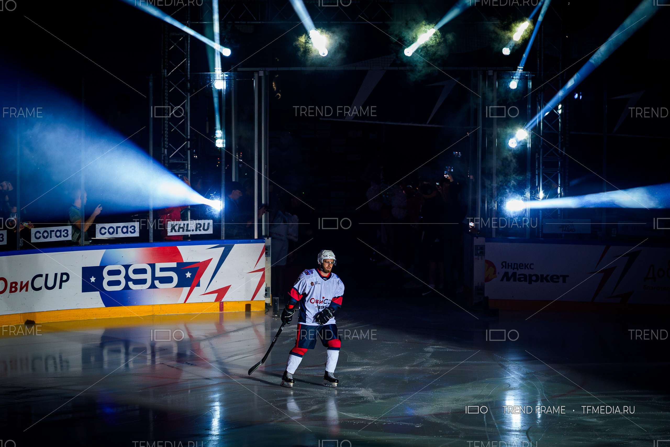 Тарас Зацепин, нападающий под номером 10, выходит на лёд в составе команды Павла Дацюка на финале Кубка Овечкина 2025 в Мытищах. Игрок в белой форме с красно-синими вставками появляется из тоннеля арены под световое шоу и аплодисменты зрителей. Премиальная спортивная фотография от TrendFrame.