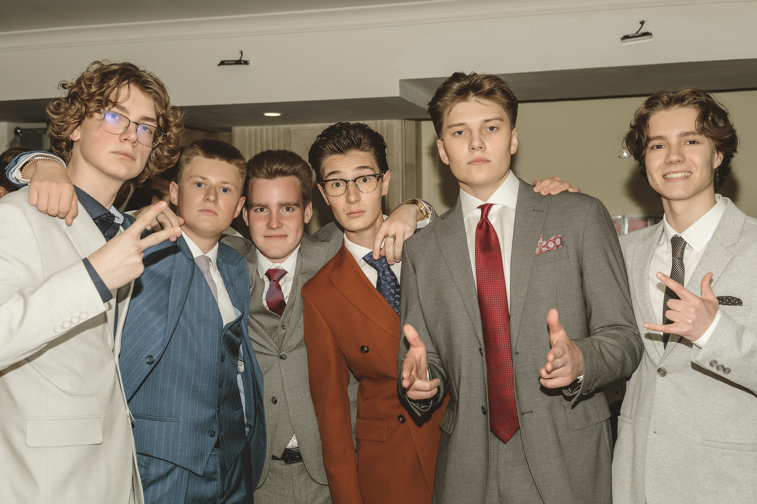 Группа из шести молодых людей, одетых в строгие костюмы, уверенно позирует в помещении. Атмосфера оживленная, с улыбками и непринужденными жестами.