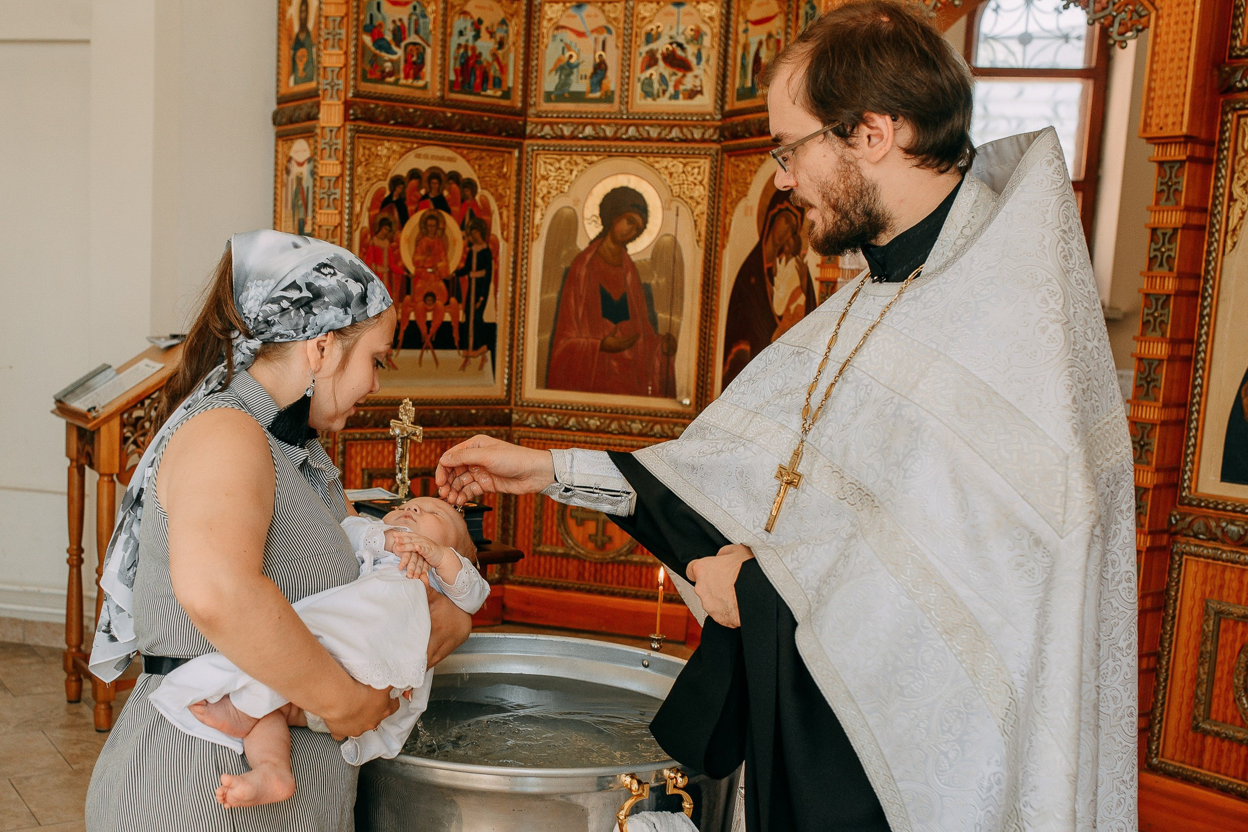 Священник совершает обряд миропомазания младенца у святой купели на фоне православного иконостаса.