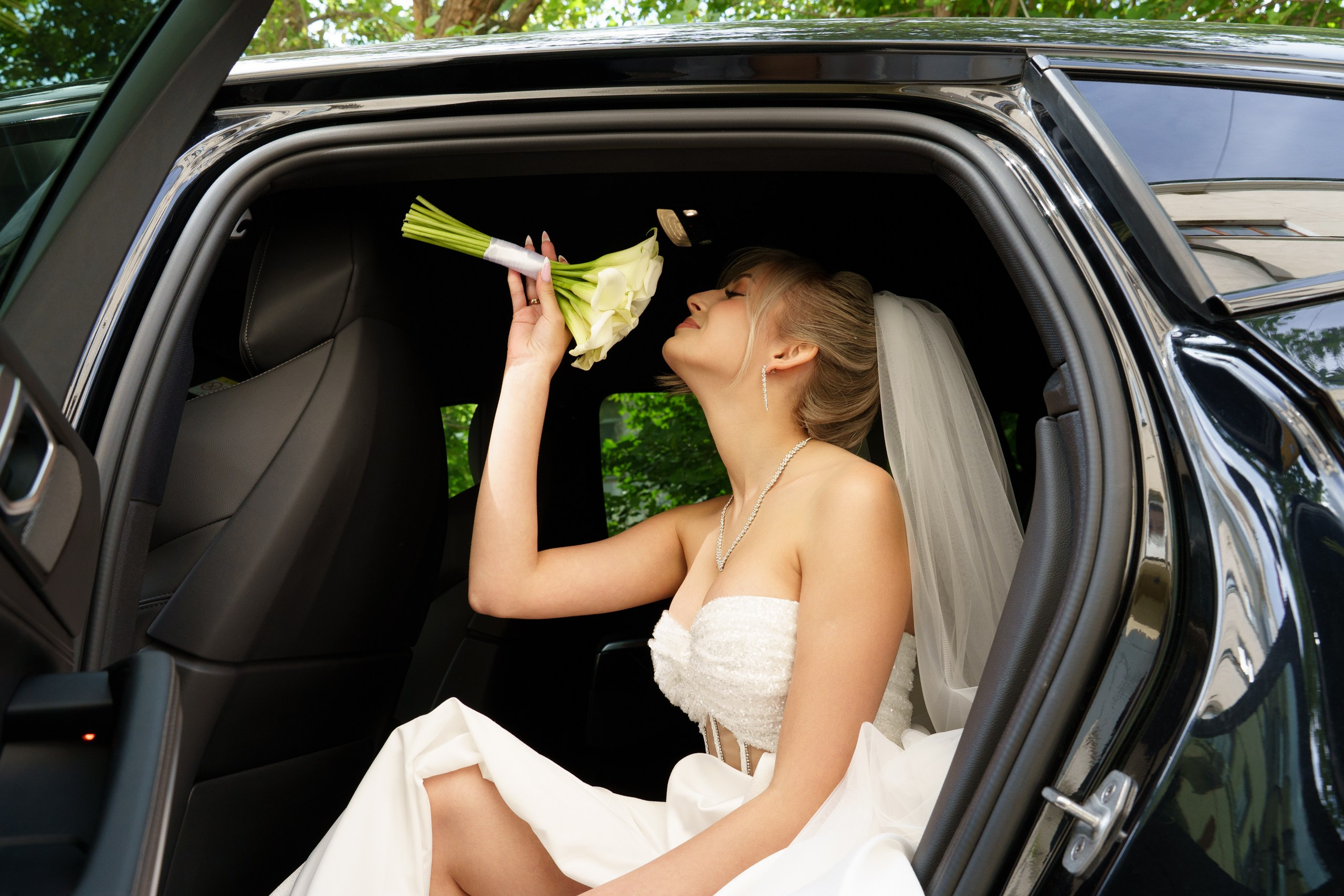 Невеста в роскошном белом платье с фатой сидит в автомобиле и вдыхает аромат своего свадебного букета. Она выглядит очень умиротворённой | свадебный фотограф с индивидуальным подходом Екатеринбург