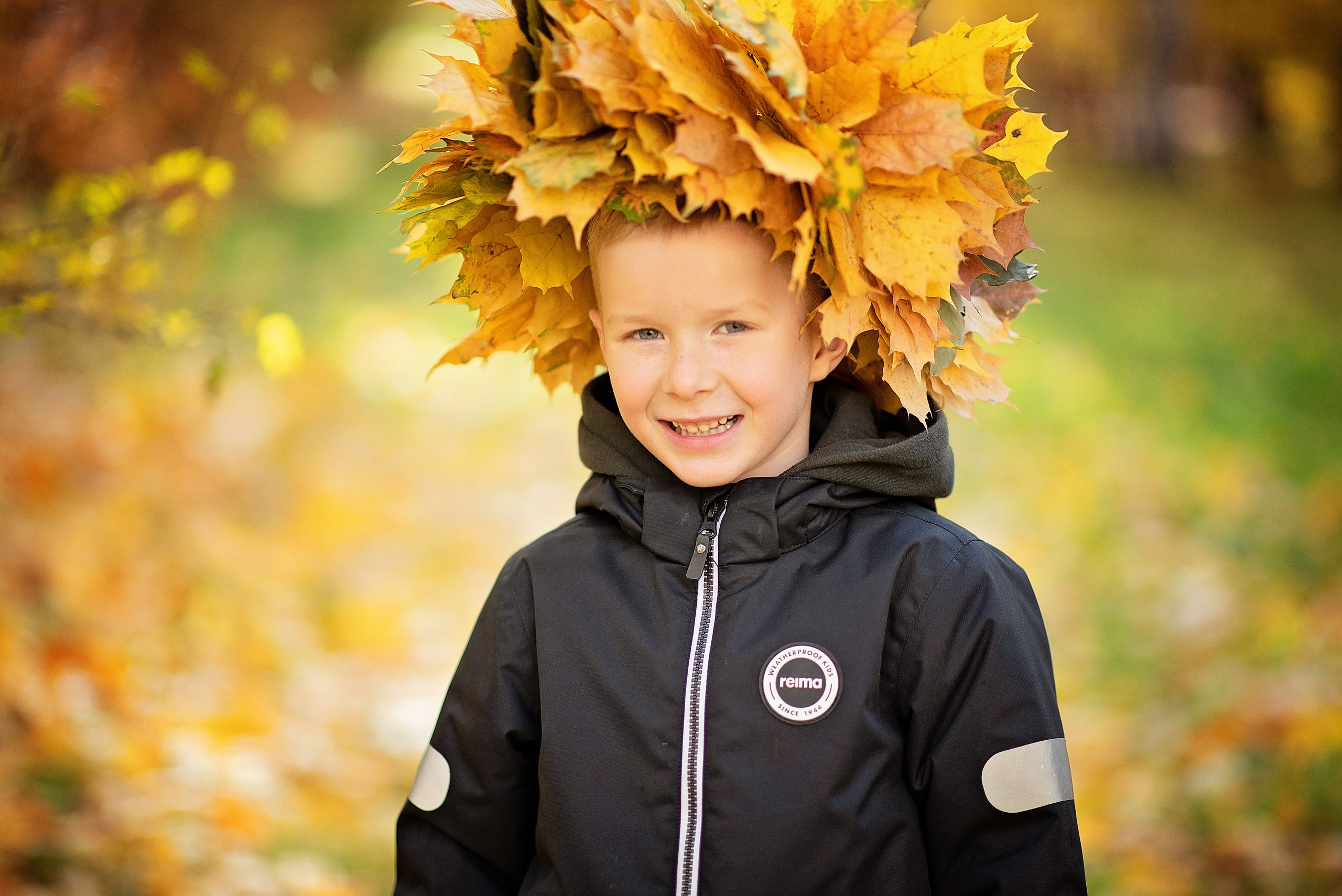мальчик стоит во дворе детского сада с венком из осенних листьев фотографируясь для выпускного альбома