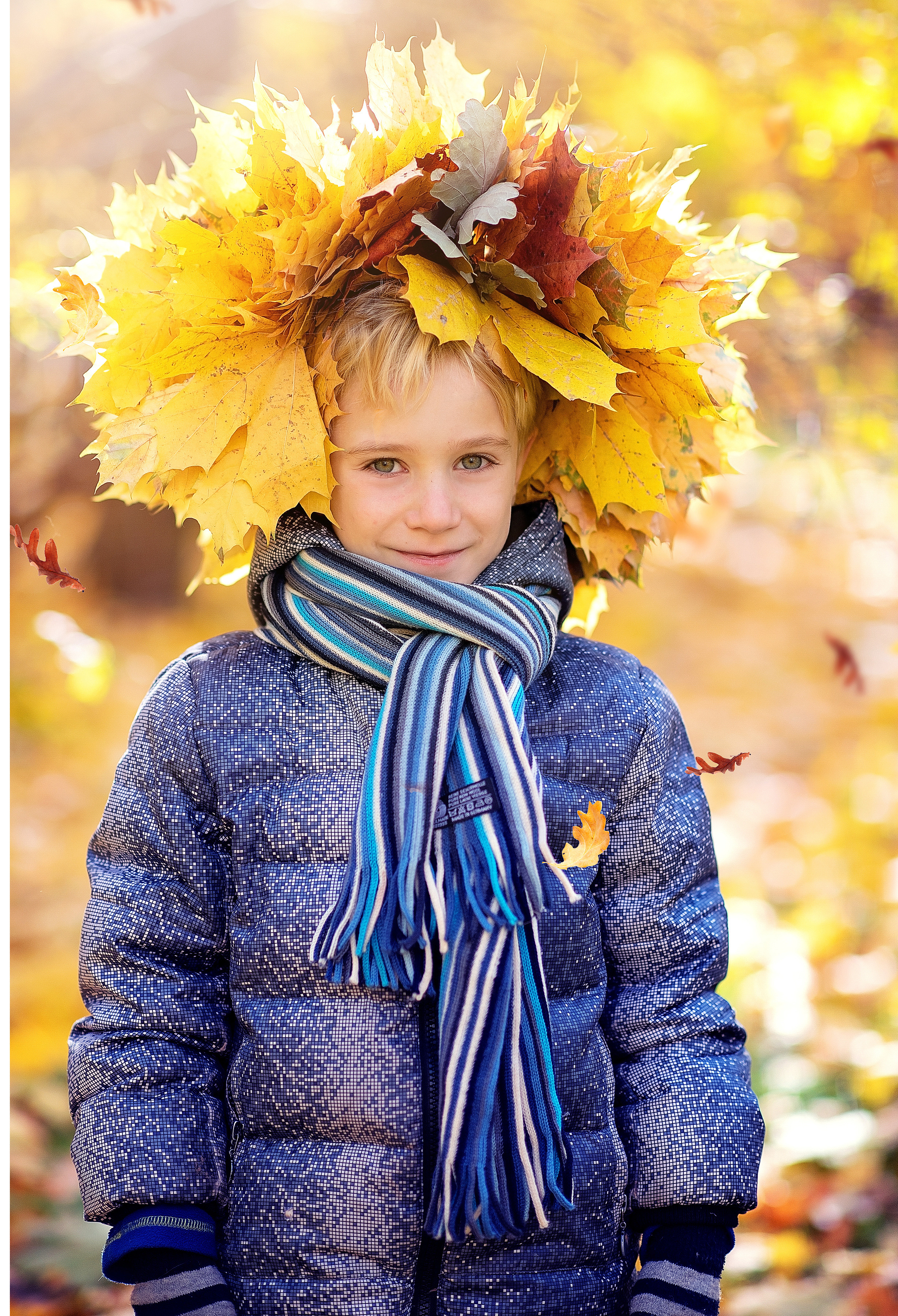 Красивый мальчик в голубой куртке и шарфике стоит во дворе детского сада с венком из осенних листьев на голове