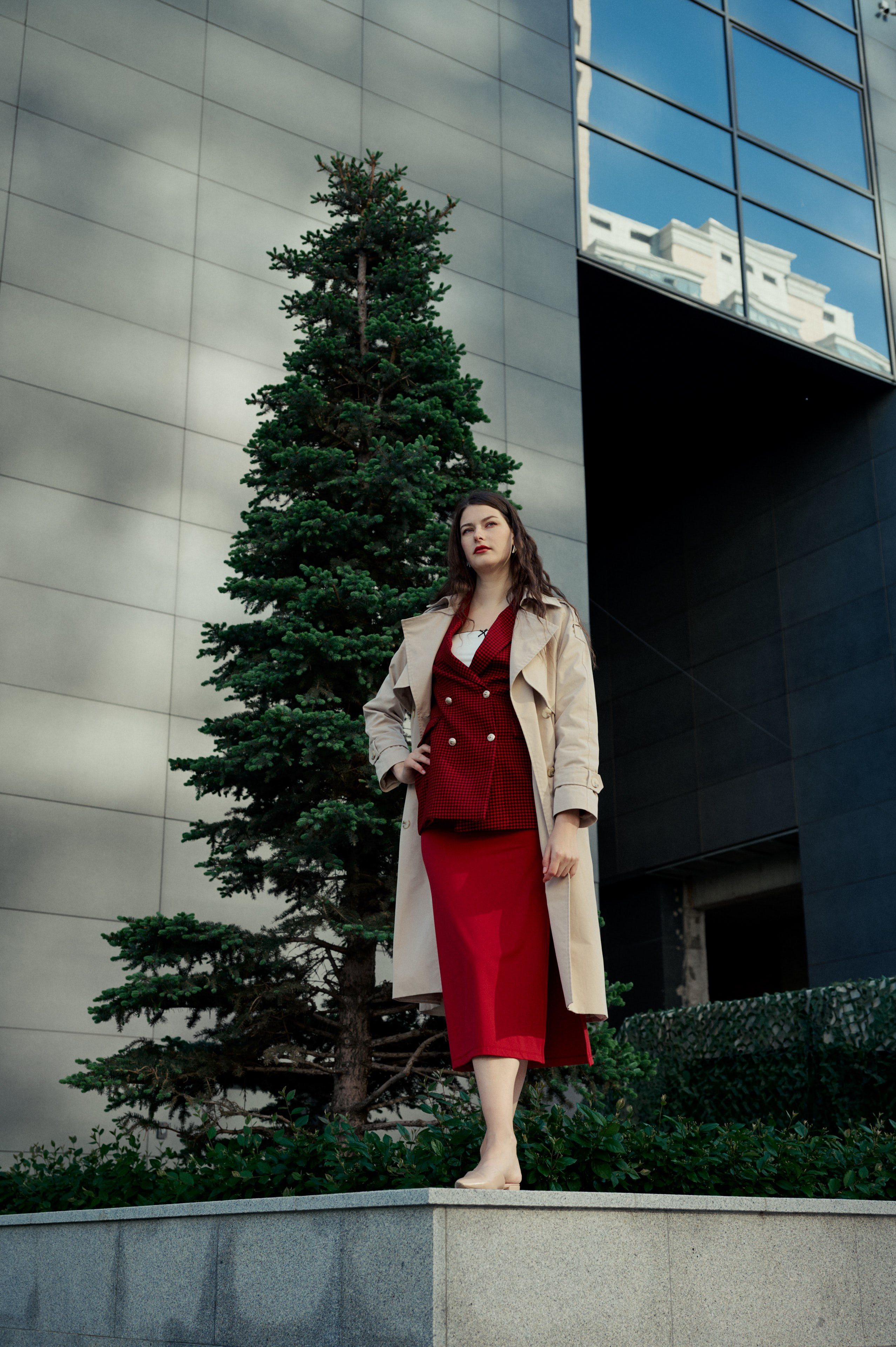 Фотосессия возле современного здания с зеркальными панелями; женщина в красном платье и светлом пальто стоит у ели.