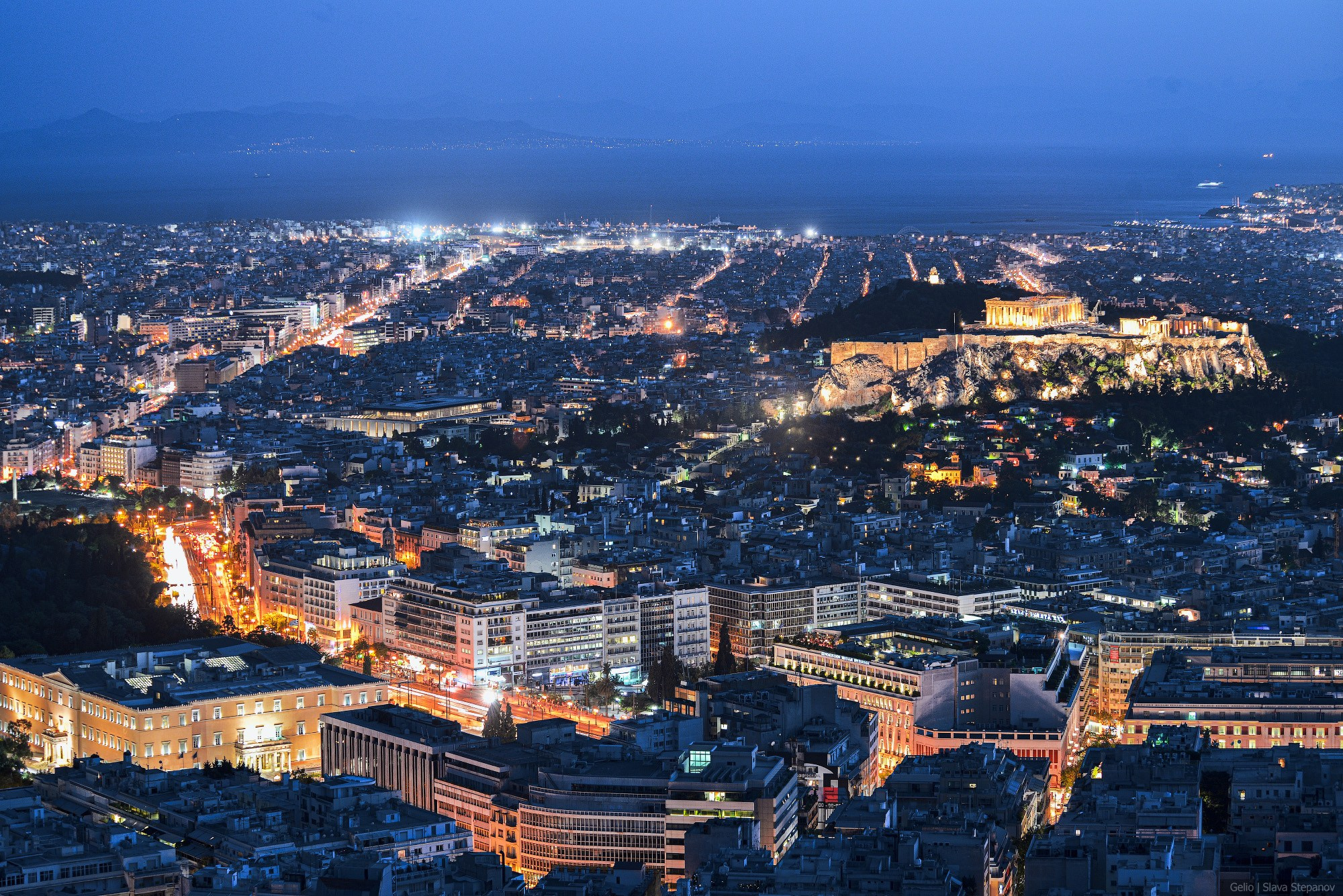 Bird's-eye view of Athens at night. The brightly illuminated historic Acropolis complex and the endless city lights stretching to the coast. Aerial photography, Slava Stepanov.
