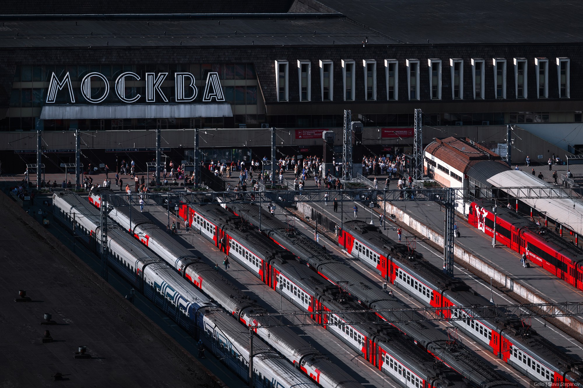 Platforms of Paveletsky railway station with a large "Moscow" facade sign, stationary passenger trains, and the flow of people. Transport photography, Gelio.