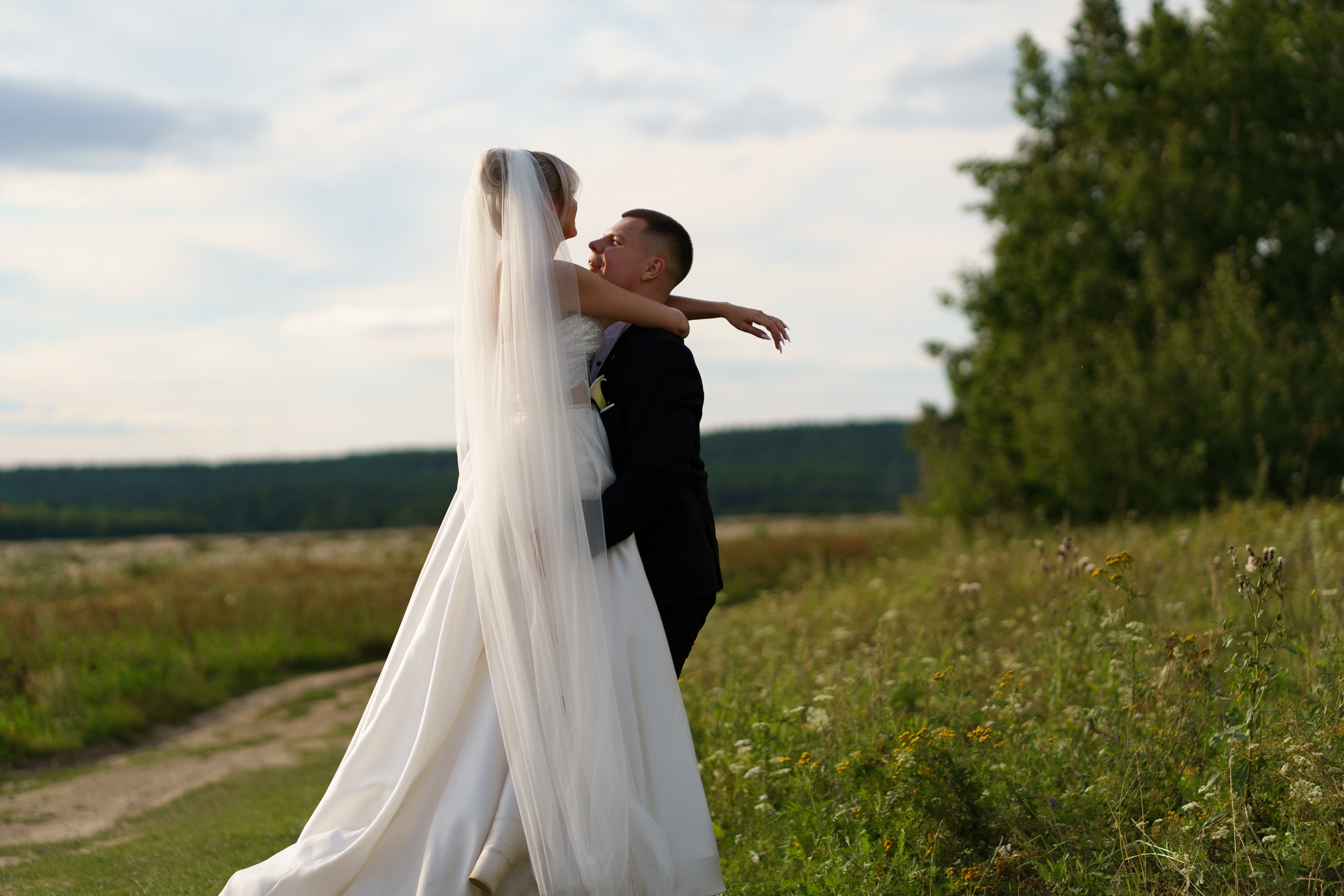 Свадебная фотосессия на открытом воздухе: невеста в пышном белом платье с фатой обнимает жениха. Вокруг — зелёные поля, высокая трава и деревья, на небе лёгкие облака |