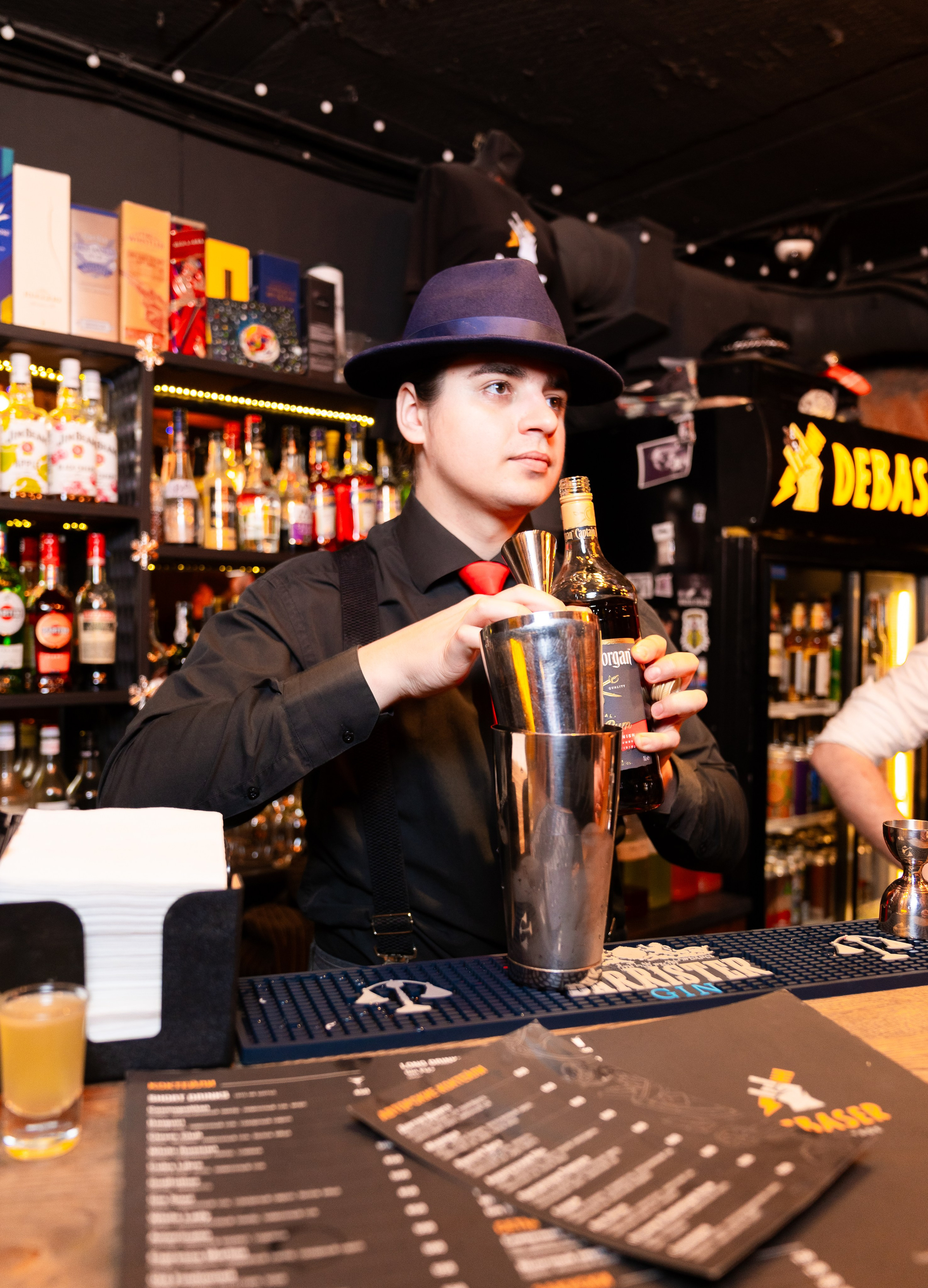 Стильный бармен в шляпе готовит коктейль с шейкером за барной стойкой в баре Debaser. Профессиональная репортажная съёмка мероприятий, фотограф Мальцев Евгений.