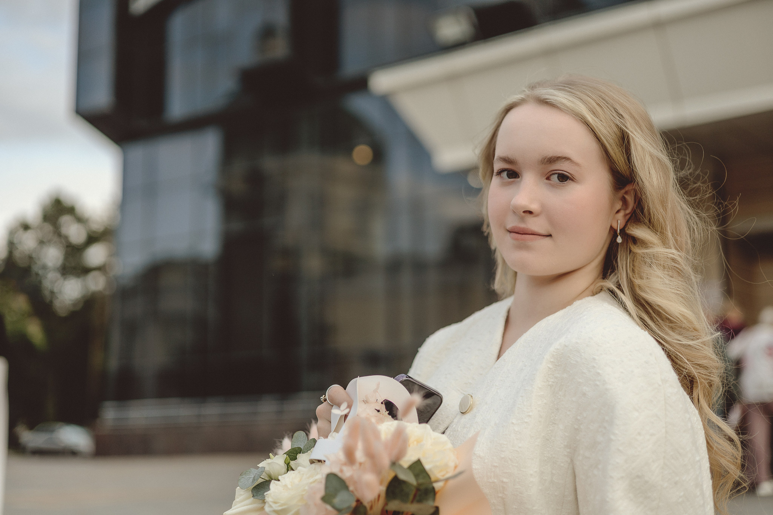 Молодая девушка с длинными светлыми волосами и нежным выражением лица держит букет кремовых роз. Она стоит перед современным стеклянным зданием, создающим атмосферу безмятежности и элегантности.