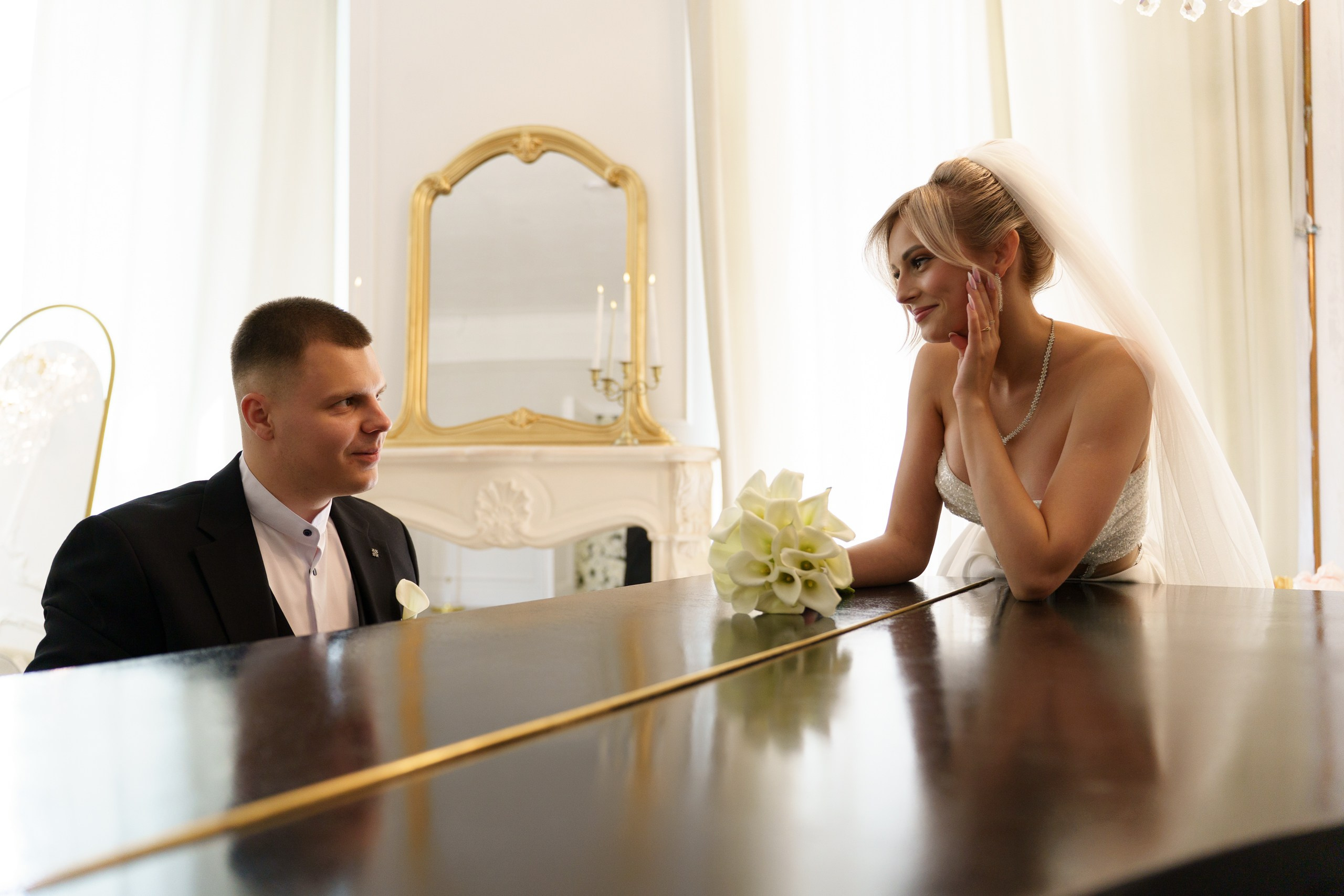 Свадебная фотосессия: жених и невеста в официальных нарядах сидят за столом. Невеста слегка наклонилась вперёд, её рука у лица, она смотрит на жениха. На столе — букет белых калл, на фоне — зеркало в золотой раме | фото для свадьбы Екатеринбург