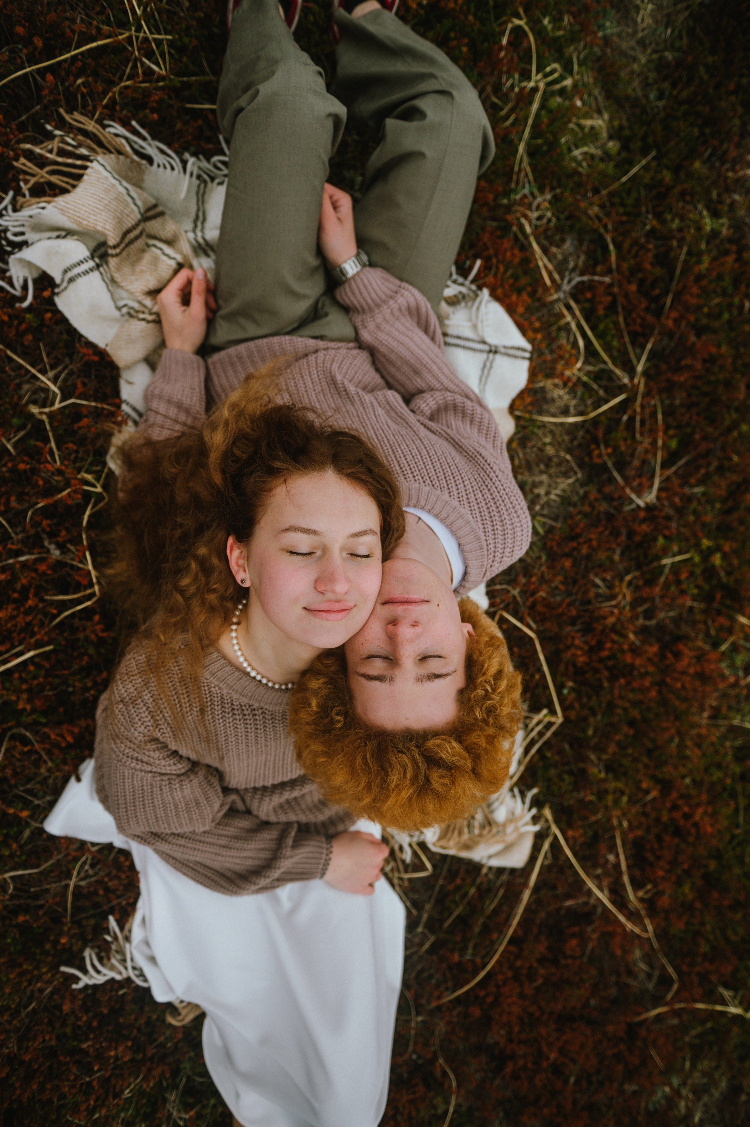Пара лежит на пледе среди травы, обнявшись. Оба с рыжими кудрями, в уютной одежде. Атмосфера спокойная, романтичная.