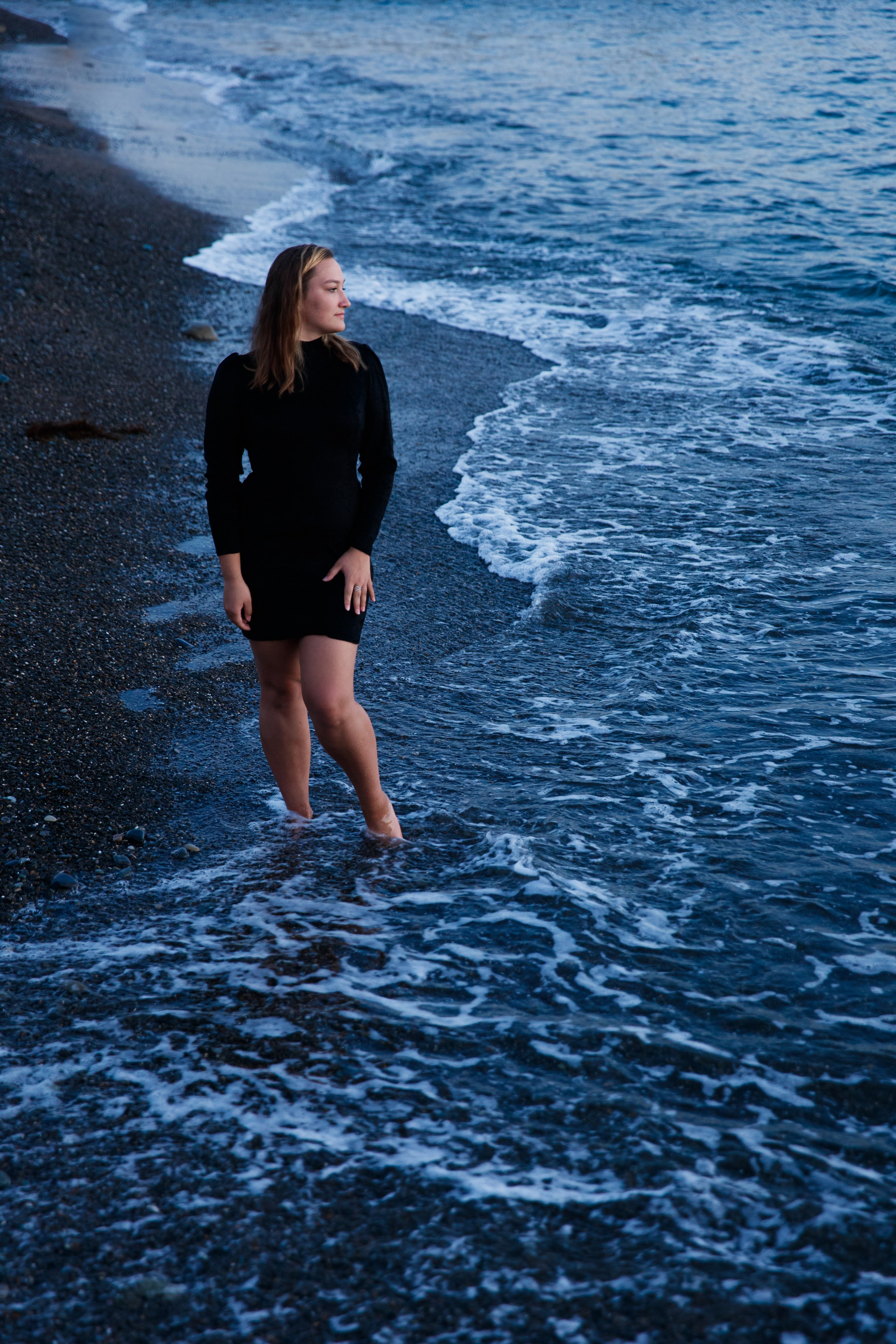 Девушка в коротком чёрном платье стоит на линии прибоя босиком