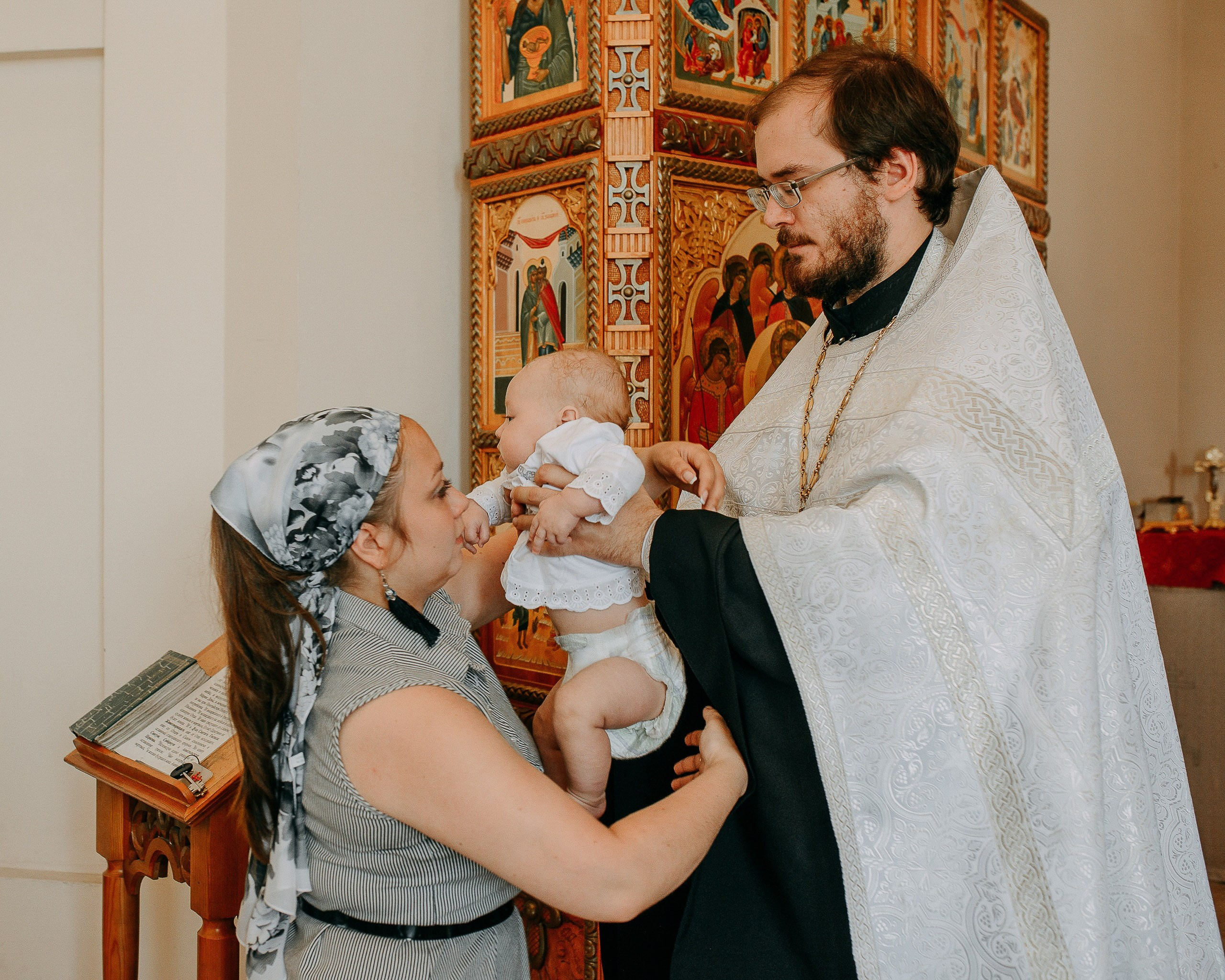 Профессиональная съемка крещения, Новосибирск: священник передает крещаемого младенца на руки крестной матери.