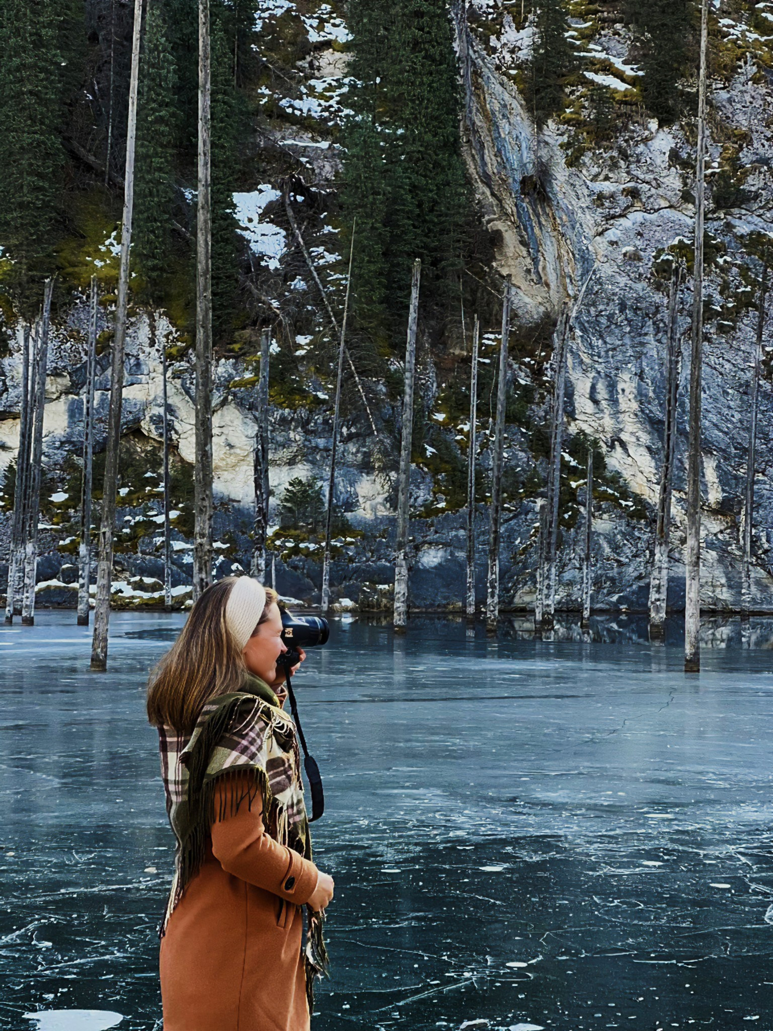 Женщина в светло-коричневом пальто с клетчатым шарфом и ободком на голове фотографирует на фотоаппарат. Она стоит на замёрзшей поверхности озера Каинды в Алматы, покрытой тонким слоем льда, за которой виднеются деревья, торчащие из воды, и заснеженные горы. Фон украшен густыми хвойными лесами и отвесными скалами, создающими драматичный зимний пейзаж.