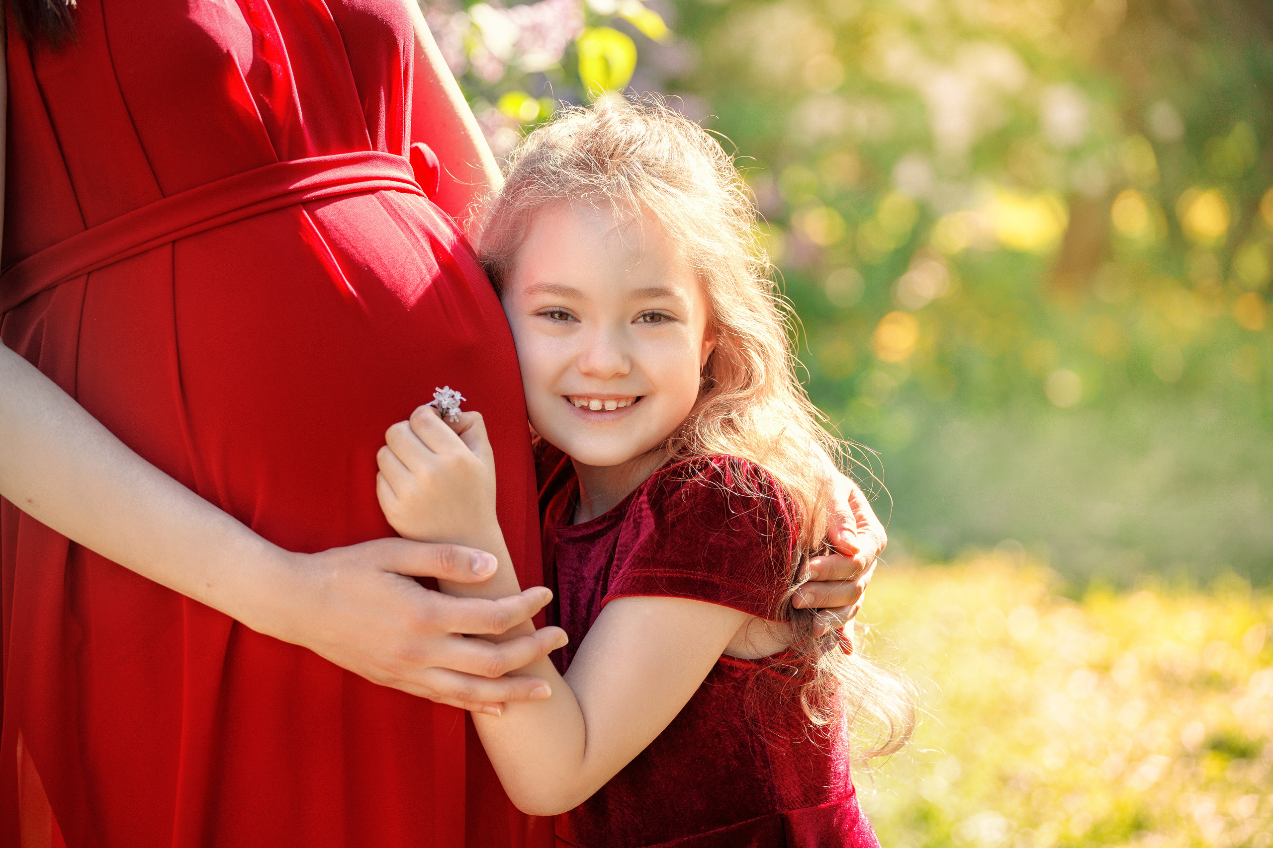 Дочка слушает беременный живот у мамы на фоне цветущих деревьев в парке