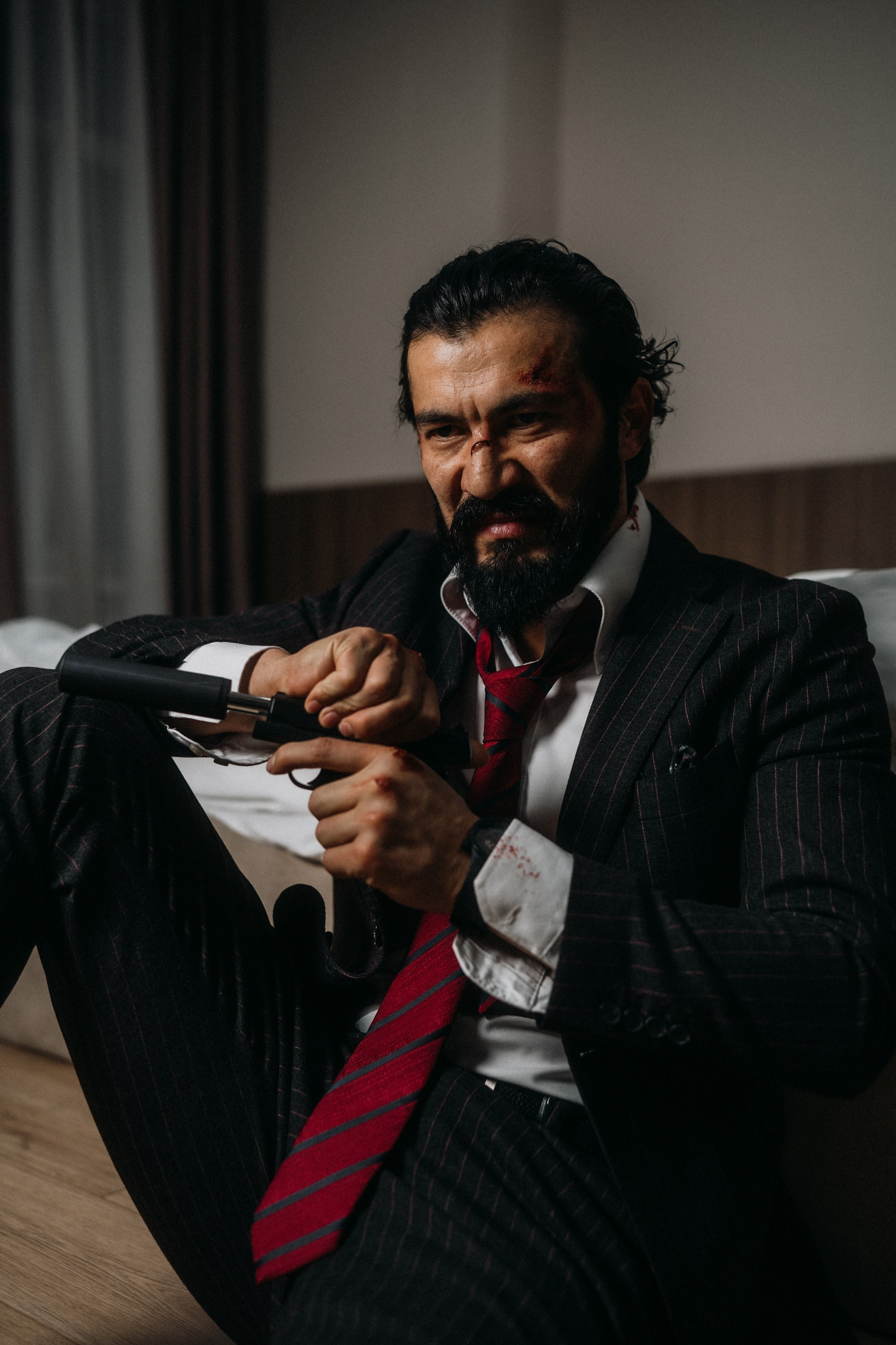 Мужчина в костюме с кровавыми ранами и пистолетом в руках сидит на полу, показывая драматический кинематографический образ, фотосессия в стиле Джон Уик