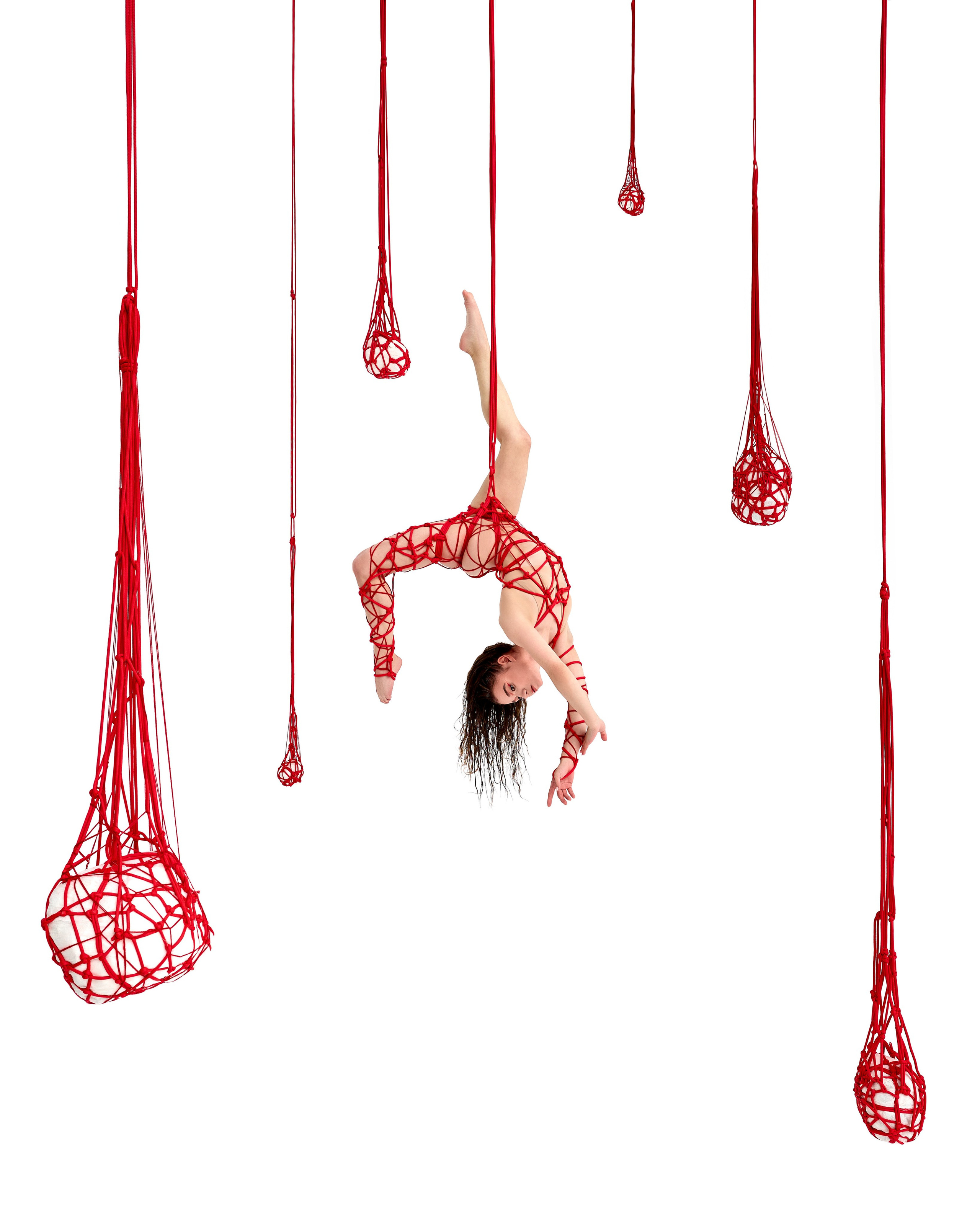 Творческий проект: на снимке изображена женщина, подвешенная в воздухе с помощью красных верёвок, которые образуют сложную сетку вокруг её тела. Она выполнена в акробатической позе, изгибаясь вниз, её ноги и руки обвиты верёвками. Вокруг неё висят несколько крупных сеток, также сделанных из красных верёвок. Белый фон подчёркивает контраст между ярким красным и элегантной позой модели