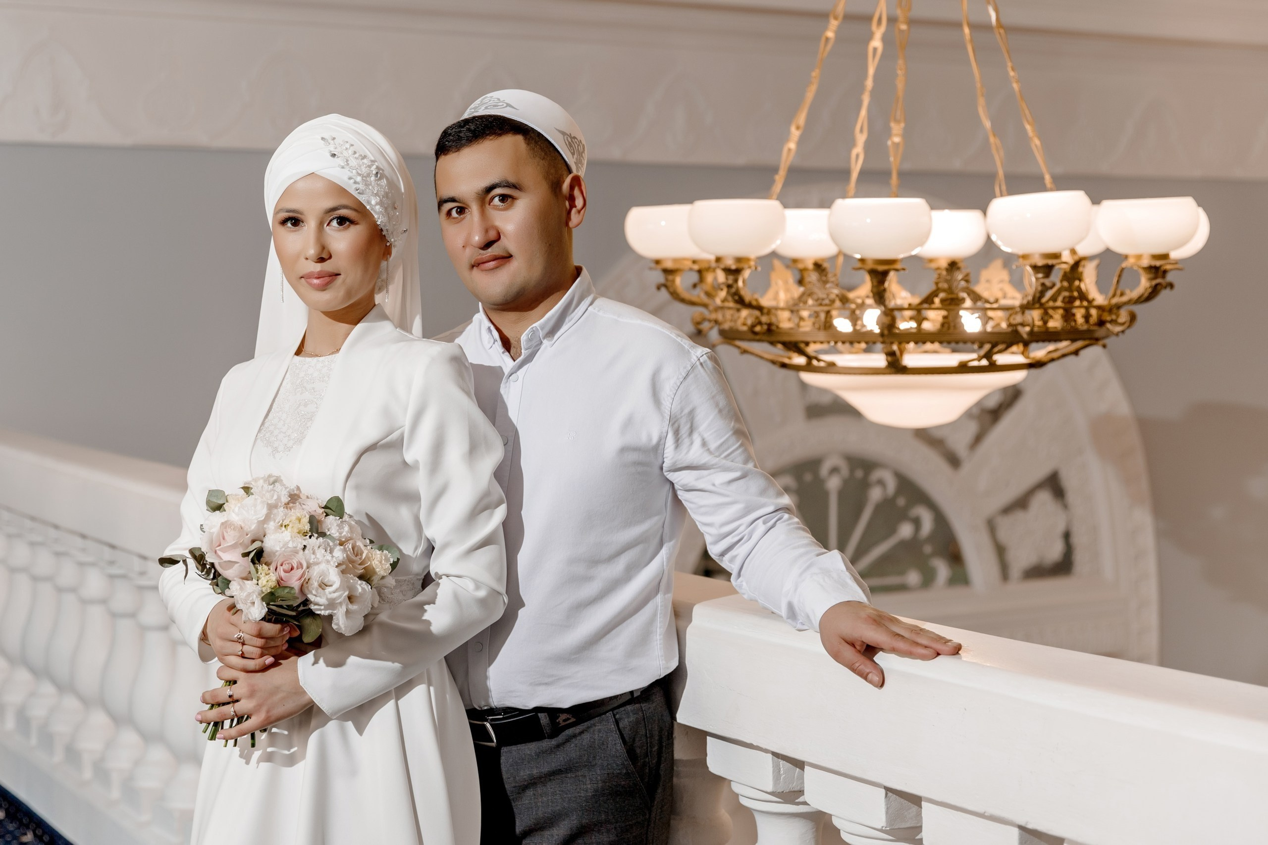 Никах. Жених и невеста в традиционных нарядах — она с букетом, он в белой тюбетейке. Стоят у балюстрады под люстрой.