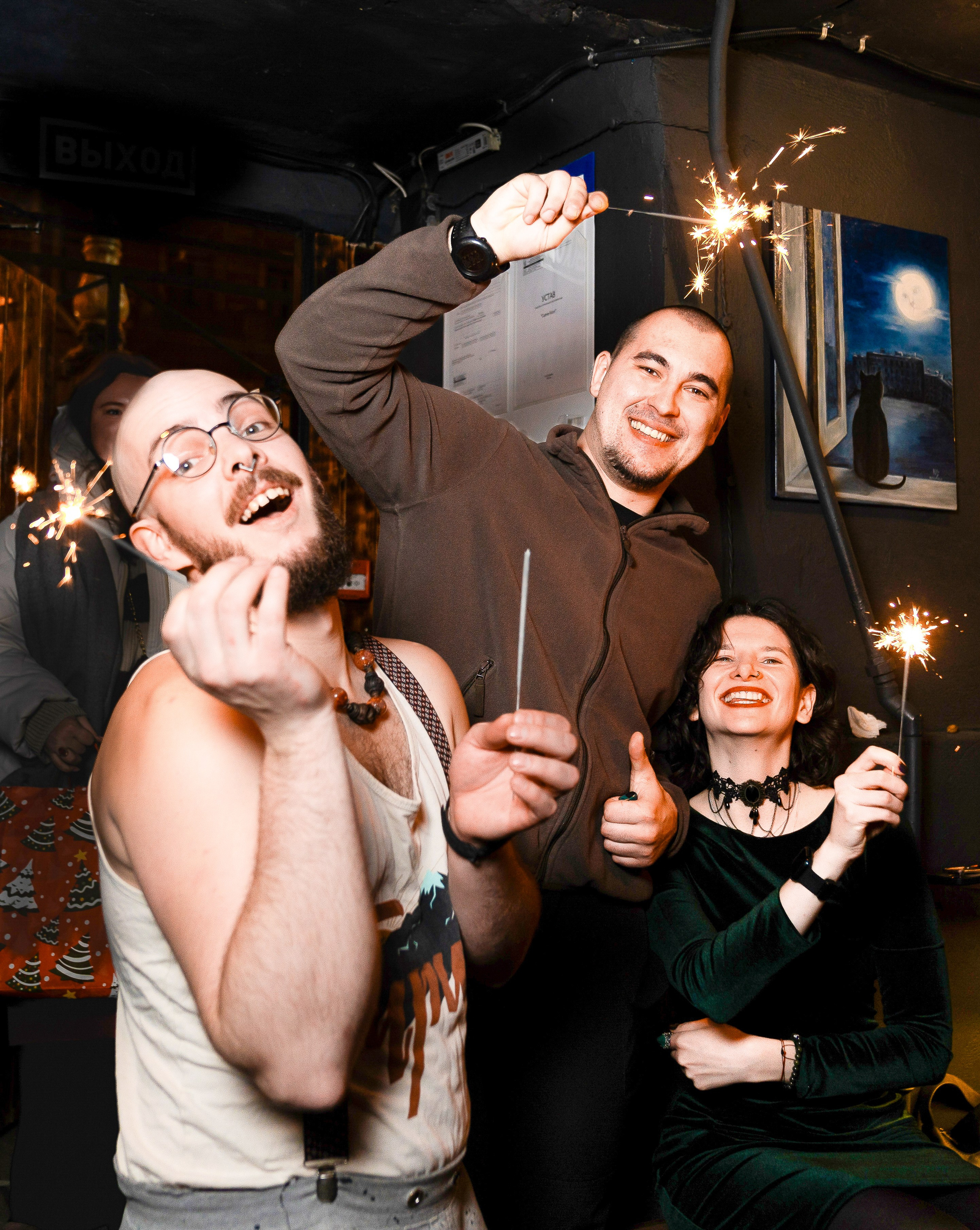 Весёлая компания с горящими бенгальскими огнями на вечеринке: парень в белой майке, мужчина в худи и улыбающаяся девушка. Праздничная репортажная фотосъёмка, фотограф Мальцев Евгений.