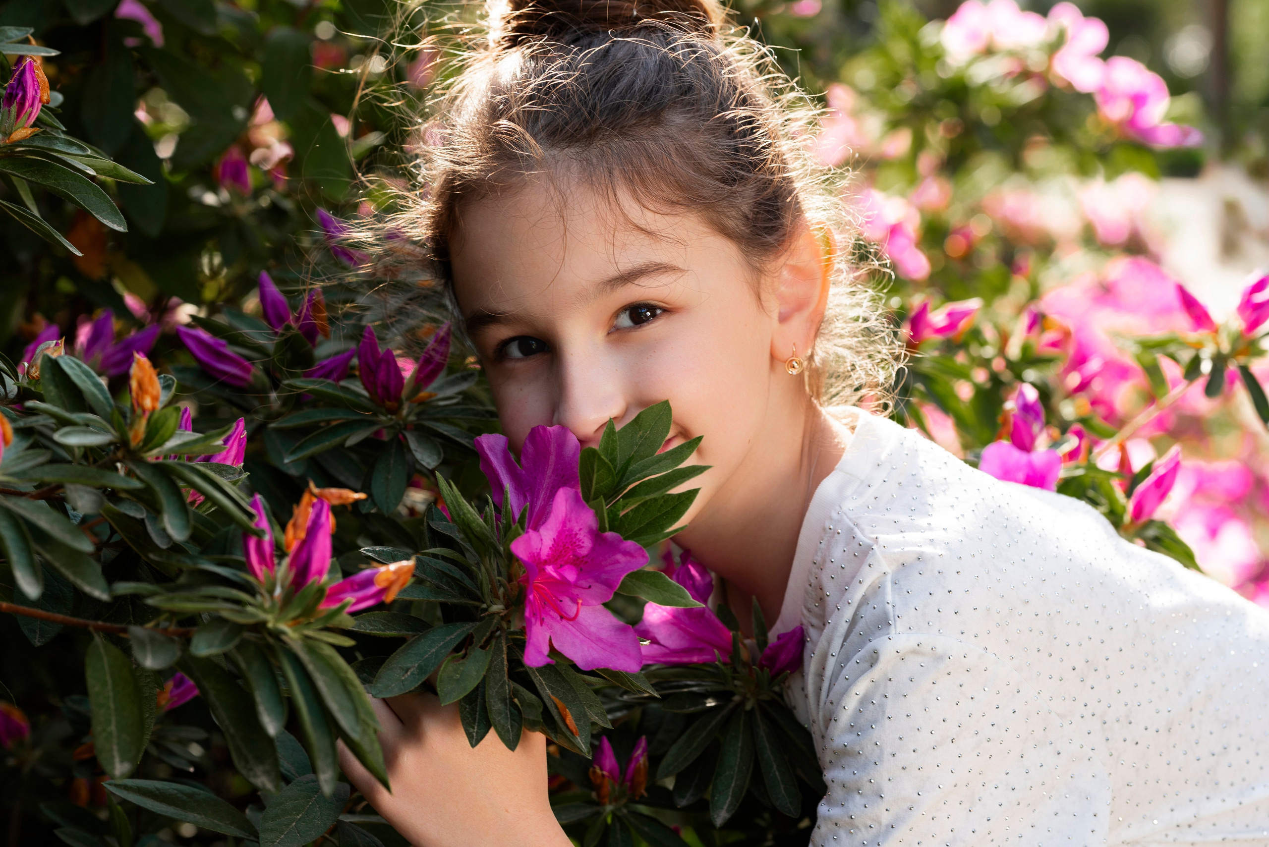 Девочка в белой одежде, обнимающий куст с ярко-розовыми рододендроном на фоне зеленых листьев, под теплыми лучами солнца, в Дендрарии Сочи