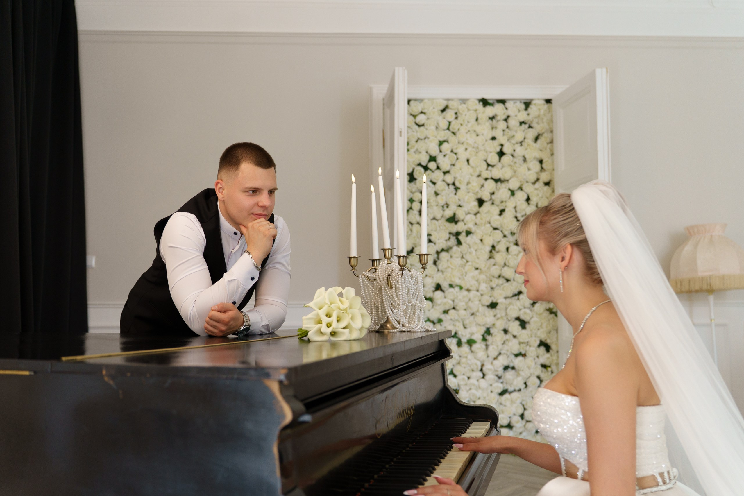 Свадебная фотосессия у рояля: жених в элегантном наряде с интересом смотрит на невесту, играющую на пианино. Невеста в изысканном белом платье с фатой погружена в музыку. Интерьер дополнен свечами, букетом калл и цветочной стеной на фоне | тематическая свадебная фотосессия Екатеринбург