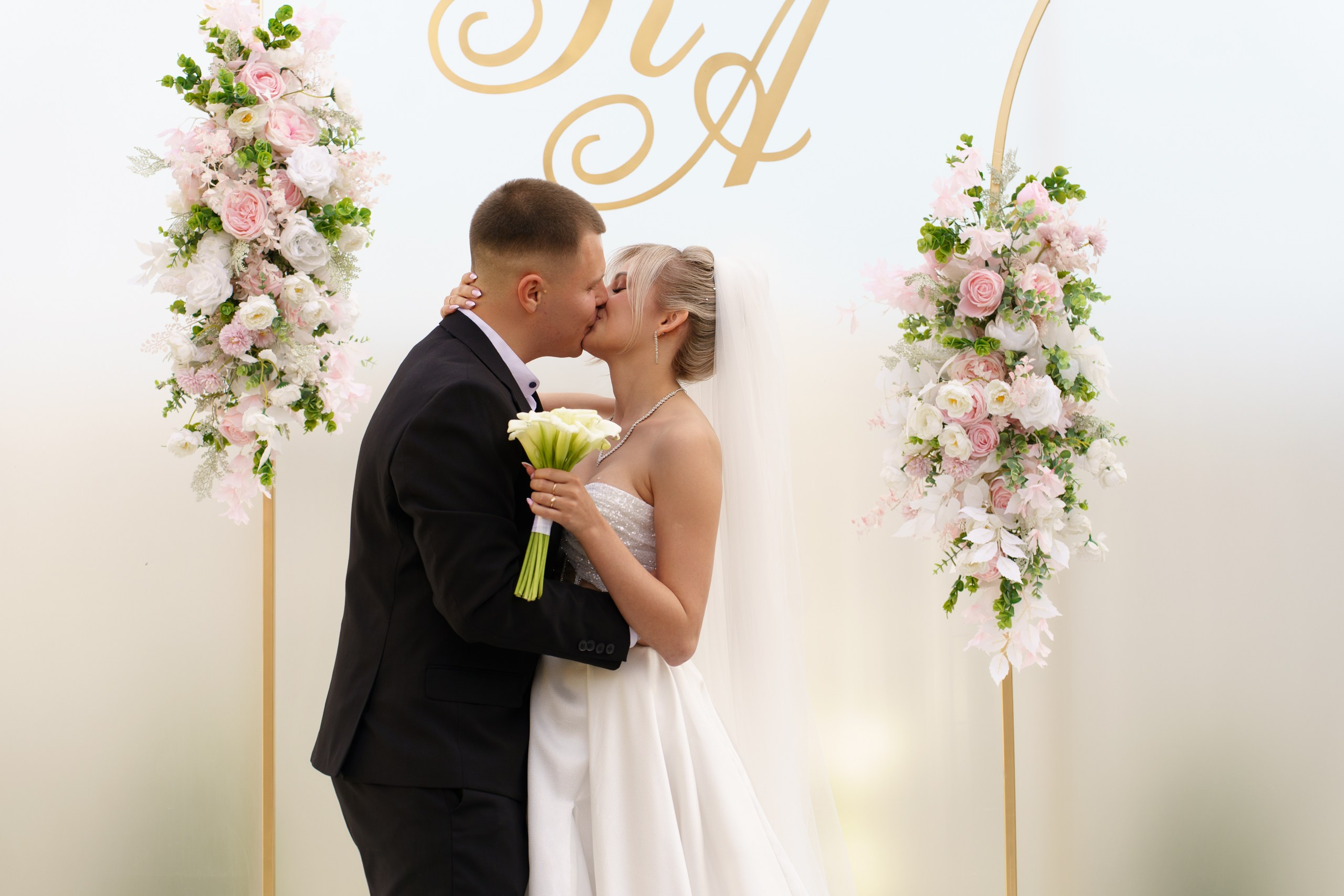 Романтический кадр со свадьбы: молодожёны в объятиях друг друга, их губы сливаются в нежном поцелуе. Цветочная композиция и монограмма на арке создают атмосферу сказочного торжества | свадебный фотограф Екатеринбург
