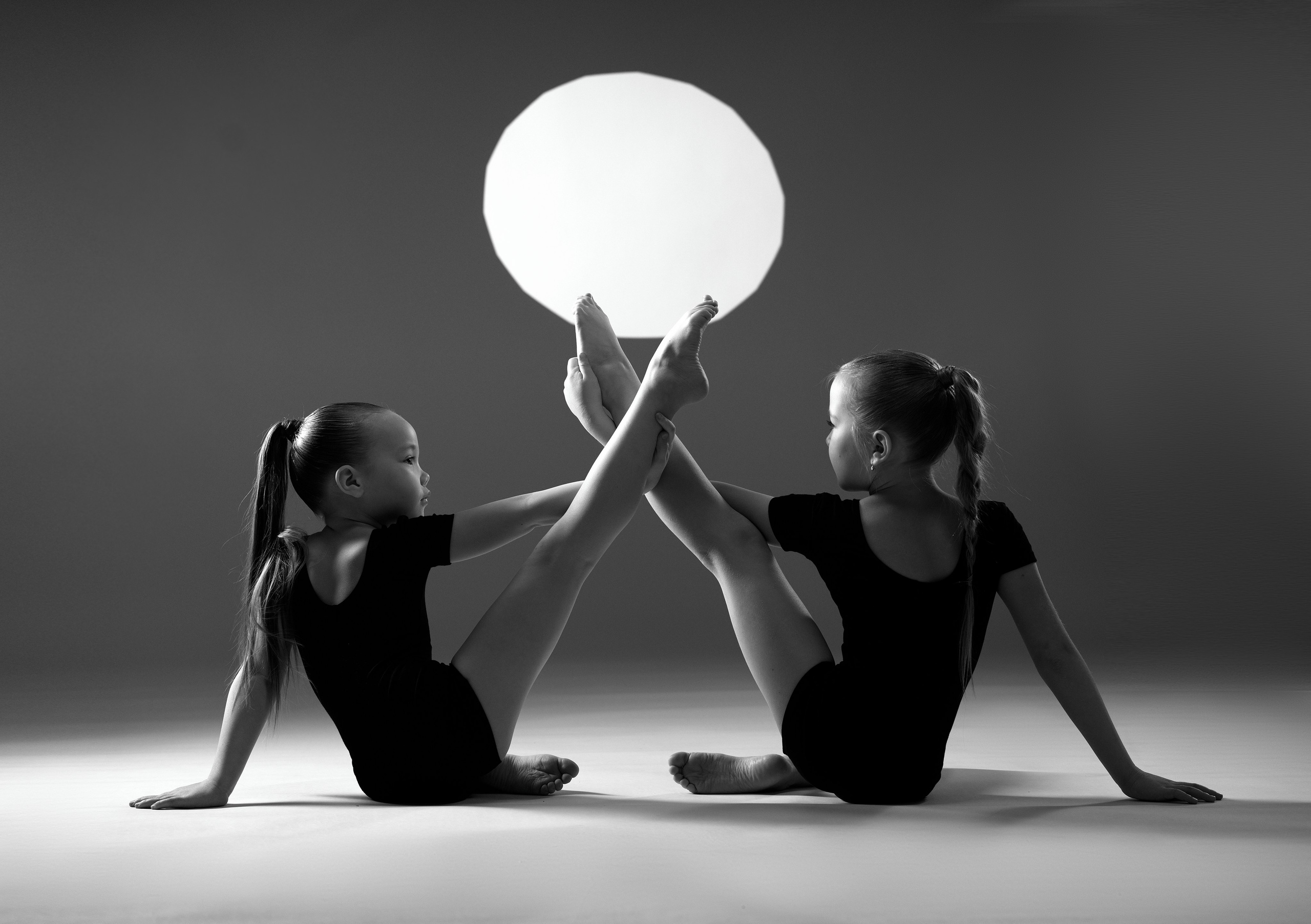 Художественная черно-белая фотография двух гимнасток в шпагате, поддерживающих светящийся шар стопами. Спортивный арт-портрет в фотостудии Тюмени.