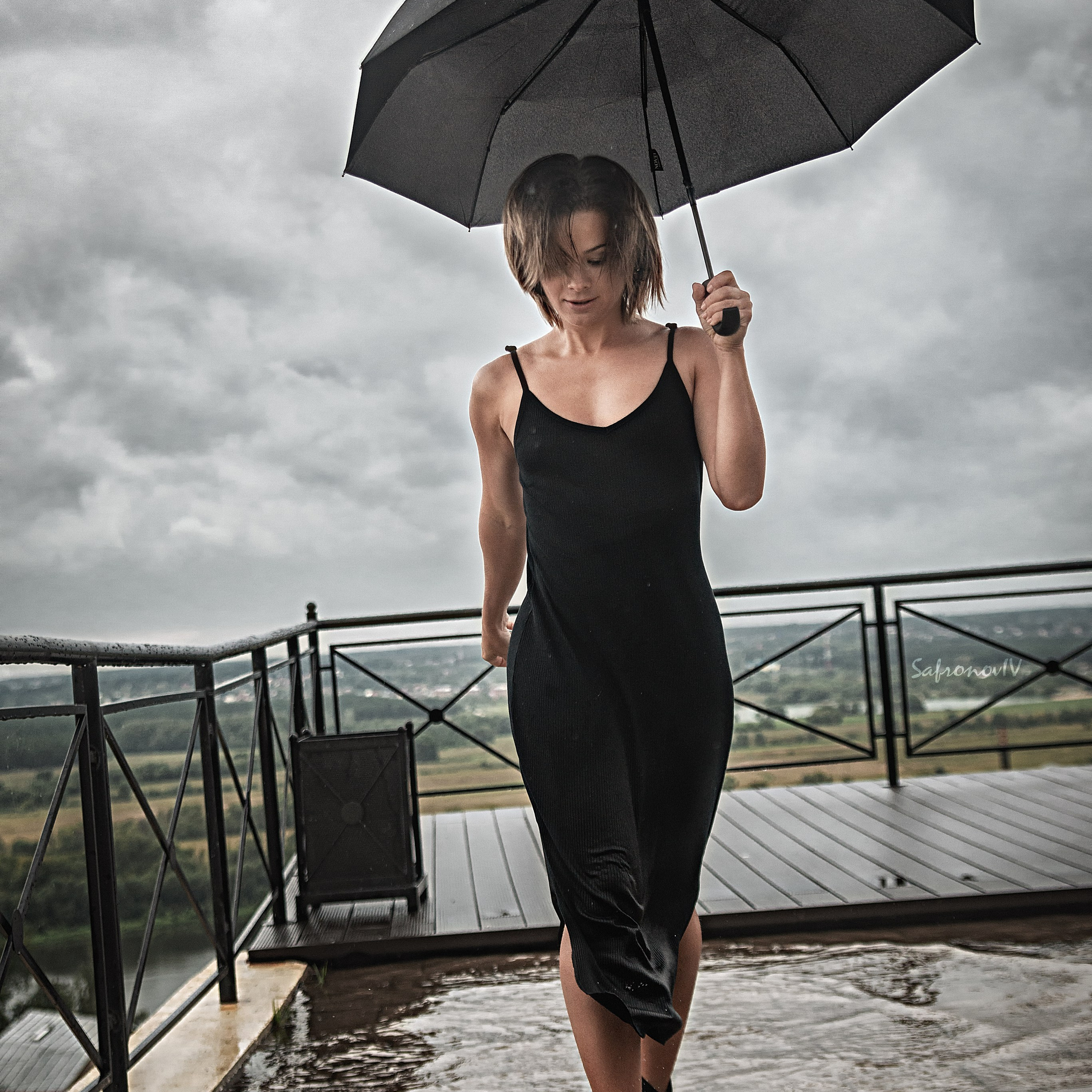На изображении запечатлен момент, полный драматизма и красоты. Женщина в черном платье уверенно шагает по мокрой террасе, держа в руках черный зонт. Ее образ подчеркивает элегантность и силу, а дождь и пасмурное небо создают атмосферу загадочности и романтики. На заднем плане открывается вид на природу, что добавляет глубину и контраст к композиции.Ключевые слова: портретный фотограф в Москве, фотограф Сафронов Иван, дождь, элегантность, природа, вдохновение.