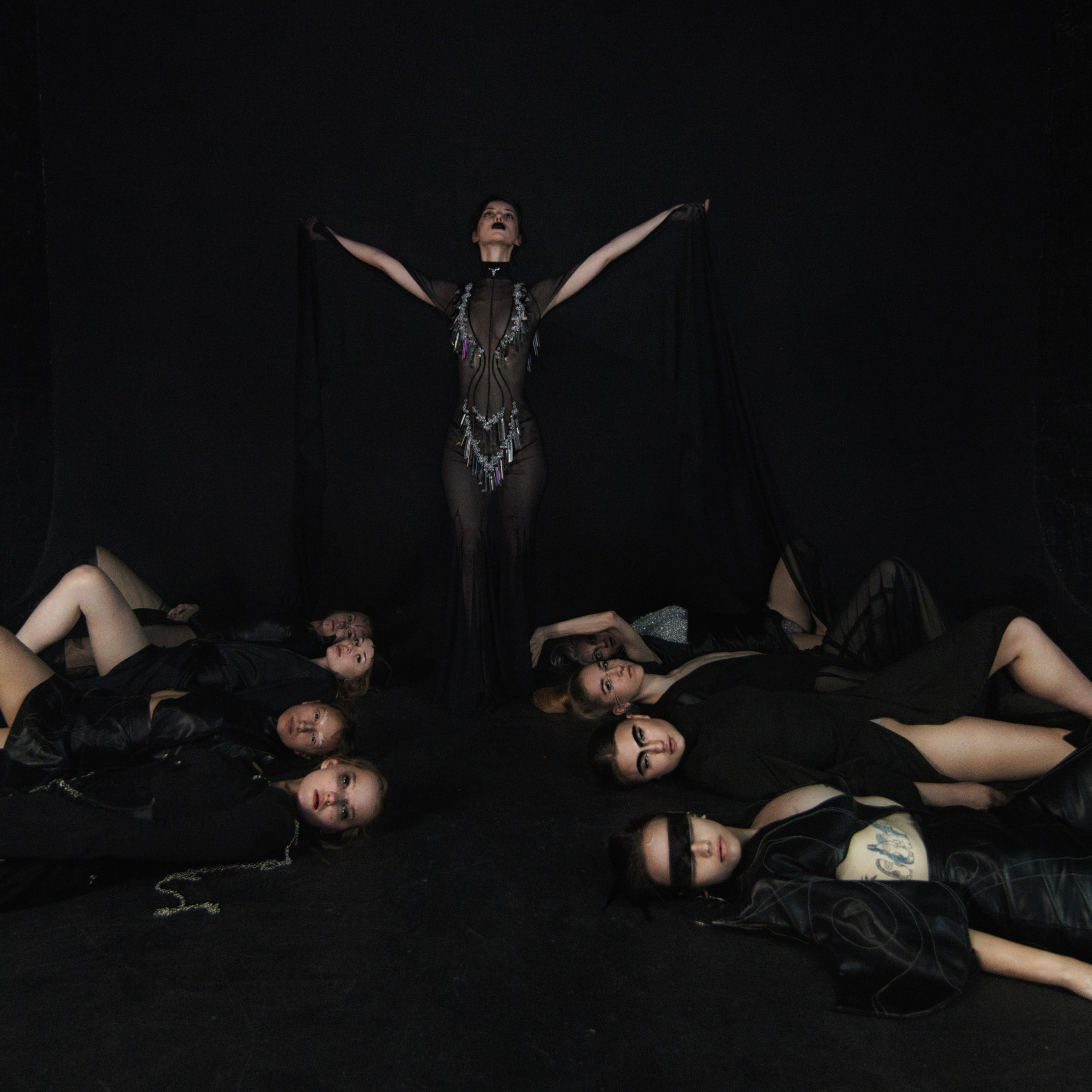 группа женщин в черных костюмах, лежащих на полу, с центральной фигурой, стоящей с поднятыми руками. Атмосфера снимка драматичная и загадочная, с акцентом на контраст между центральной фигурой и остальными. Фон темный, что подчеркивает выразительность поз и костюмов.Заголовок: «Танец теней: сила и единство в черном»Ключевые слова: танцевальные костюмы, сценические выступления, драматичная фотосессия, черный стиль, портретный фотограф, Москва, фотограф Сафронов Иван.