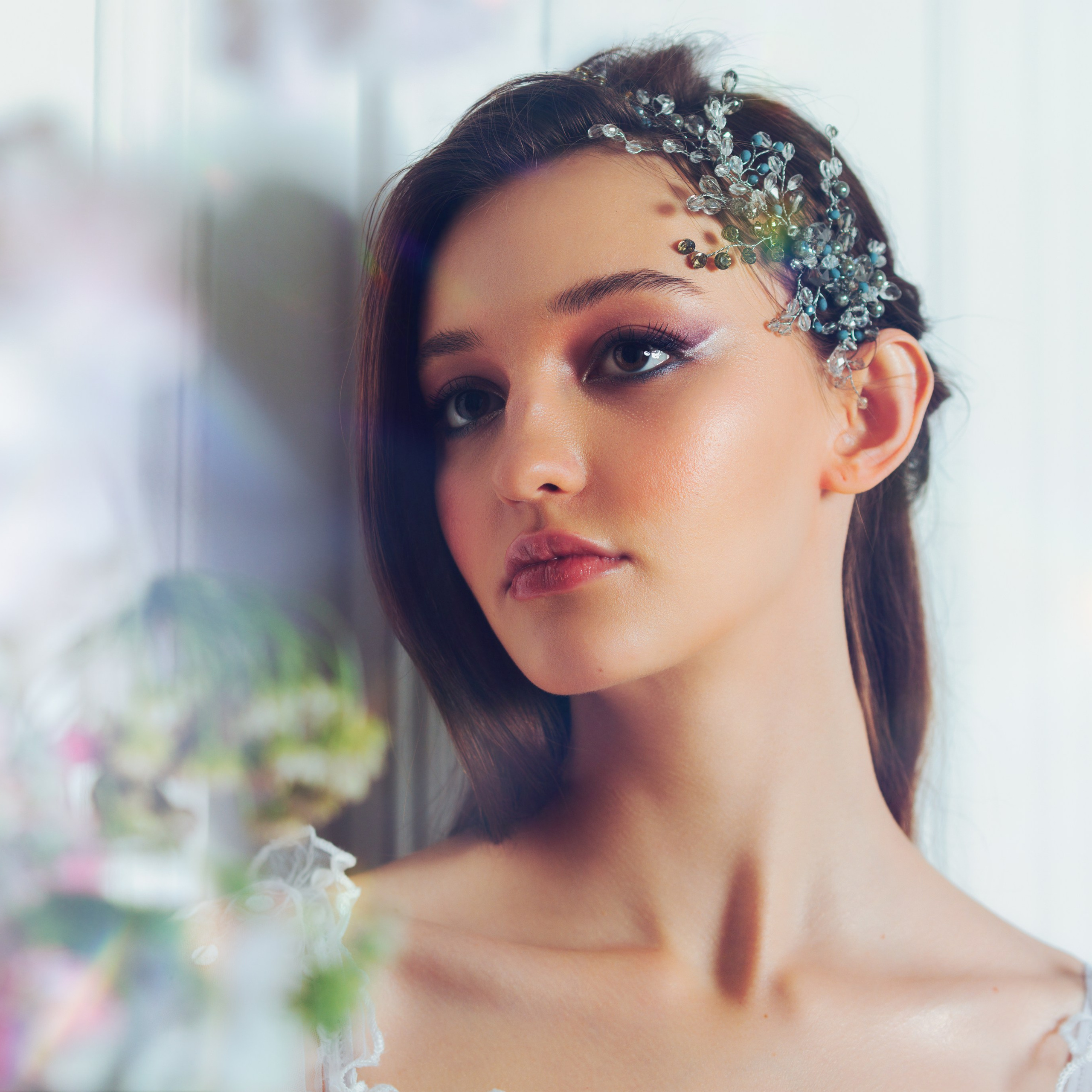 молодая девушка с утонченным макияжем и элегантной прической. На ее волосах красуется изысканное украшение с кристаллами, добавляющее образу роскоши. Фон размытый, с элементами зелени и света, что создает атмосферу легкости и романтики. Девушка смотрит вдаль, создавая ощущение задумчивости и мечтательности.Заголовок: «Элегантность в деталях: свадебный образ»Ключевые слова: свадебный макияж, украшение для волос, романтический образ, портрет, фотосессия, фотограф Сафронов Иван, Москва.