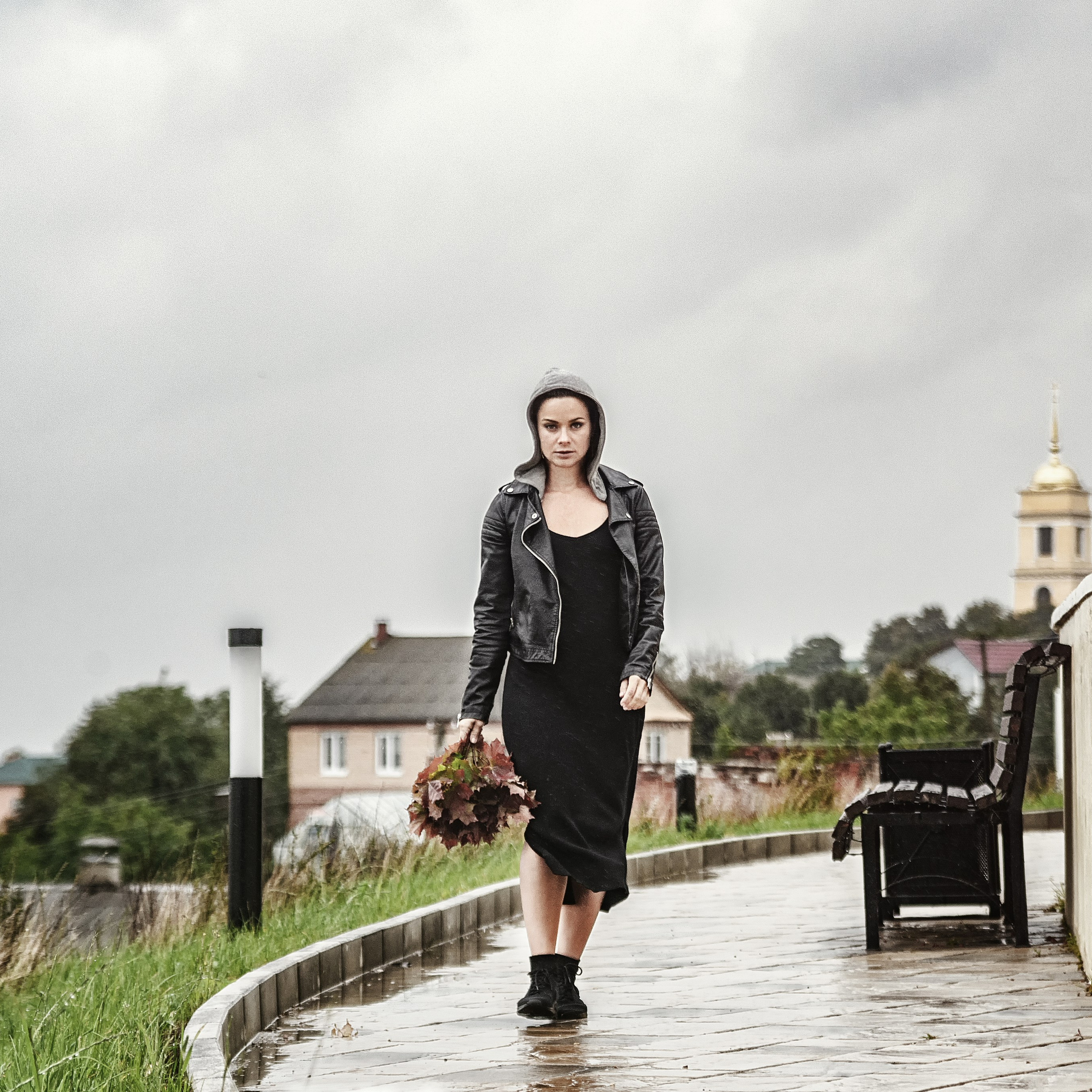 На фотографии изображена девушка, идущая по мокрой дорожке в дождливый день. Она одета в стильное черное платье и кожаную куртку, что придает образу нотку гранжа. В руках у нее букет из осенних листьев, добавляющий тепла в прохладную атмосферу. На заднем плане видны дома и церковь с золотым куполом, создающие уютный фон. Слева от девушки стоит кресло из искусственного ротанга, а справа — белая стена.Заголовок: «Осенний гранж: прогулка под дождем»Ключевые слова: фотосессия, осенний стиль, гранж, дождь, кожаное платье, осенние листья, уличная съемка, портрет, фотограф Сафронов Иван.
