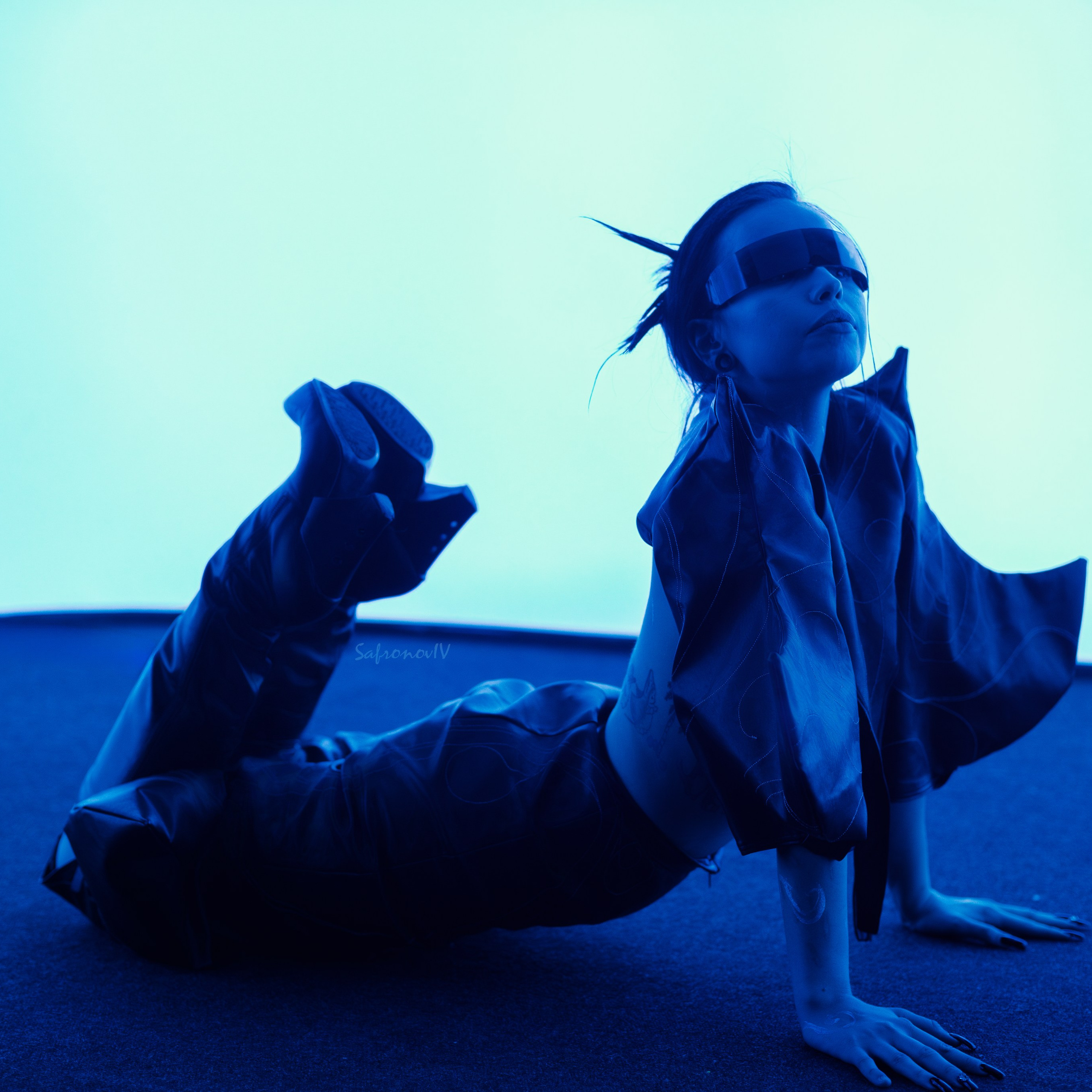 На изображении запечатлен стильный и драматичный момент. Человек лежит на полу, опираясь на руки, в расслабленной позе. Его образ дополнен блестящими черными кожаными брюками и объемной рубашкой, которая развевается, создавая ощущение движения. На голове закреплены очки виртуальной реальности, что добавляет футуристичности. Свет, окрашенный в холодный голубой оттенок, подчеркивает текстуры и создает атмосферу загадочности.**Ключевые слова:** портретный фотограф в Москве, фотограф Сафронов Иван, стиль, мода, футуризм, драматический свет, кожа, виртуальная реальность.