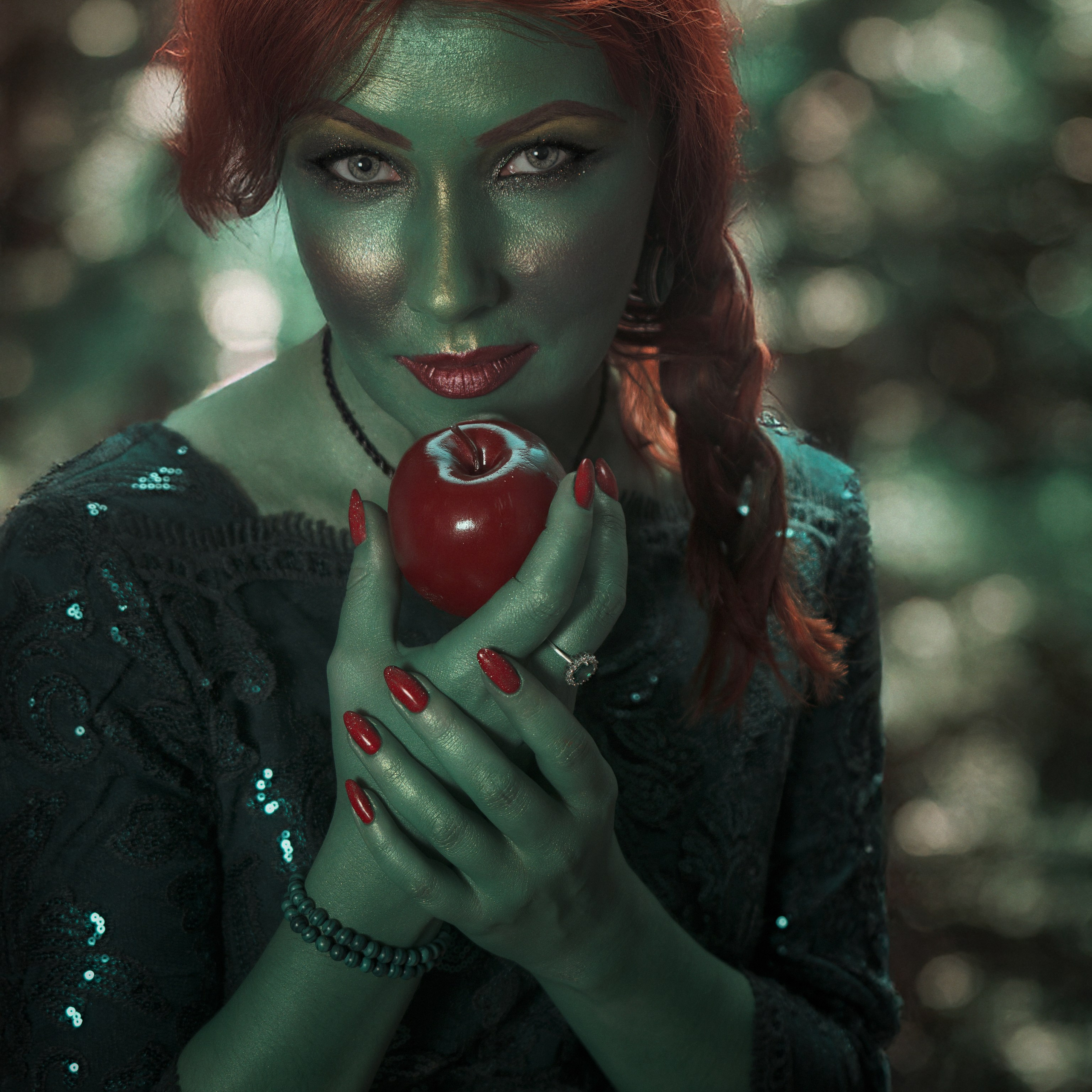 На изображении представлена женщина с ярко-рыжими волосами, одетая в тёмное платье с блёстками. Её кожа окрашена в зелёный цвет, что придаёт ей загадочный и мистический вид. В руках она держит красное яблоко, которое контрастирует с её зелёным лицом и тёмным фоном. Её макияж подчёркивает выразительность глаз и губ, а длинные ногти окрашены в красный цвет, гармонирующий с яблоком. Фон размытый, с зелёными оттенками, что усиливает атмосферу волшебства.Заголовок: «Яблоко соблазна: магия рыжей ведьмы»Ключевые слова: портретный фотограф в Москве, фотограф Сафронов Иван.