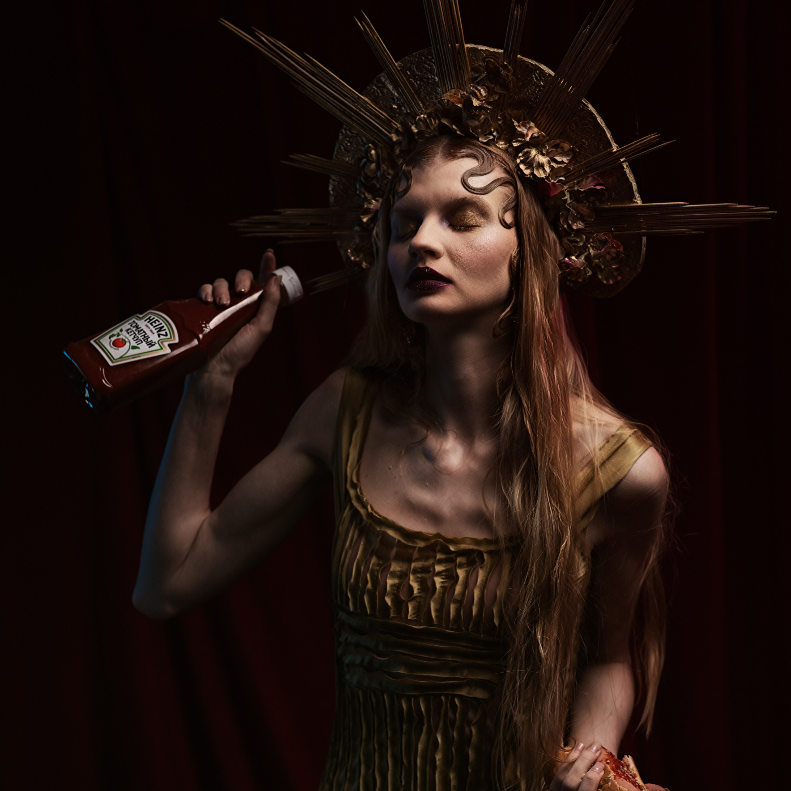 женщина в готическом стиле, с короной, напоминающей солнечные лучи, и в платье, выполненном в античном стиле. В руках она держит бутылку кетчупа Heinz и хот-дог, что добавляет образу сюрреалистичности. Освещение подчеркивает драматичность сцены, создавая атмосферу загадочности и театральности.Заголовок: «Королева кетчупа: готический стиль в современной интерпретации»Ключевые слова: фотосессия, готический стиль, корона, кетчуп, хот-дог, сюрреализм, портрет, фэнтези.