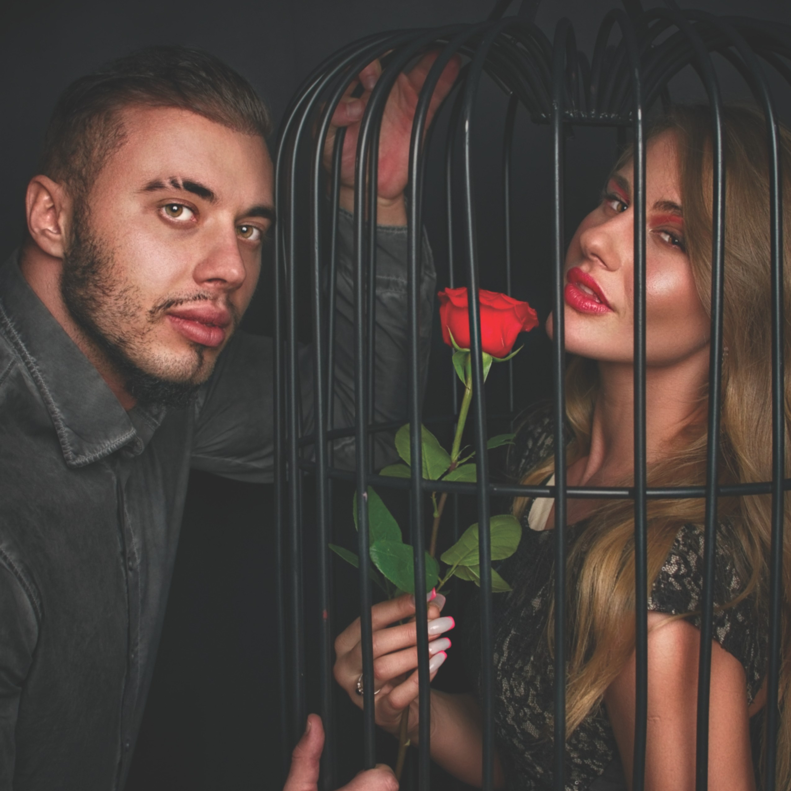 мужчина и женщина. Женщина находится внутри большой декоративной клетки, держит в руках ярко-красную розу, её взгляд направлен в сторону камеры. У неё светлые волосы, выразительный макияж с акцентом на губы и глаза, она одета в тёмное платье с узором. Мужчина стоит снаружи клетки, слегка наклонившись вперёд, его поза динамична и экспрессивна. У него короткие тёмные волосы, аккуратная борода и стильная тёмная рубашка. Его взгляд также устремлён в камеру, создавая ощущение напряжённого диалога с зрителем.Композиция построена на контрасте: клетка символизирует ограничение, а роза в руках женщины — страсть и свободу. Тёмный фон подчёркивает драматизм сцены и акцентирует внимание на моделях и реквизите. В верхней части изображения размещён логотип журнала «LOOKER MAGAZINE», в правом нижнем углу — номер страницы (25).Заголовок:«В клетке страсти: игра контрастов»Ключевые слова: портрет, студийная съёмка, модельная фотосессия, клетка, роза, драма, контраст, экспрессия, портретный фотограф, Москва, фотограф Сафронов Иван, реквизит, художественная съёмка.