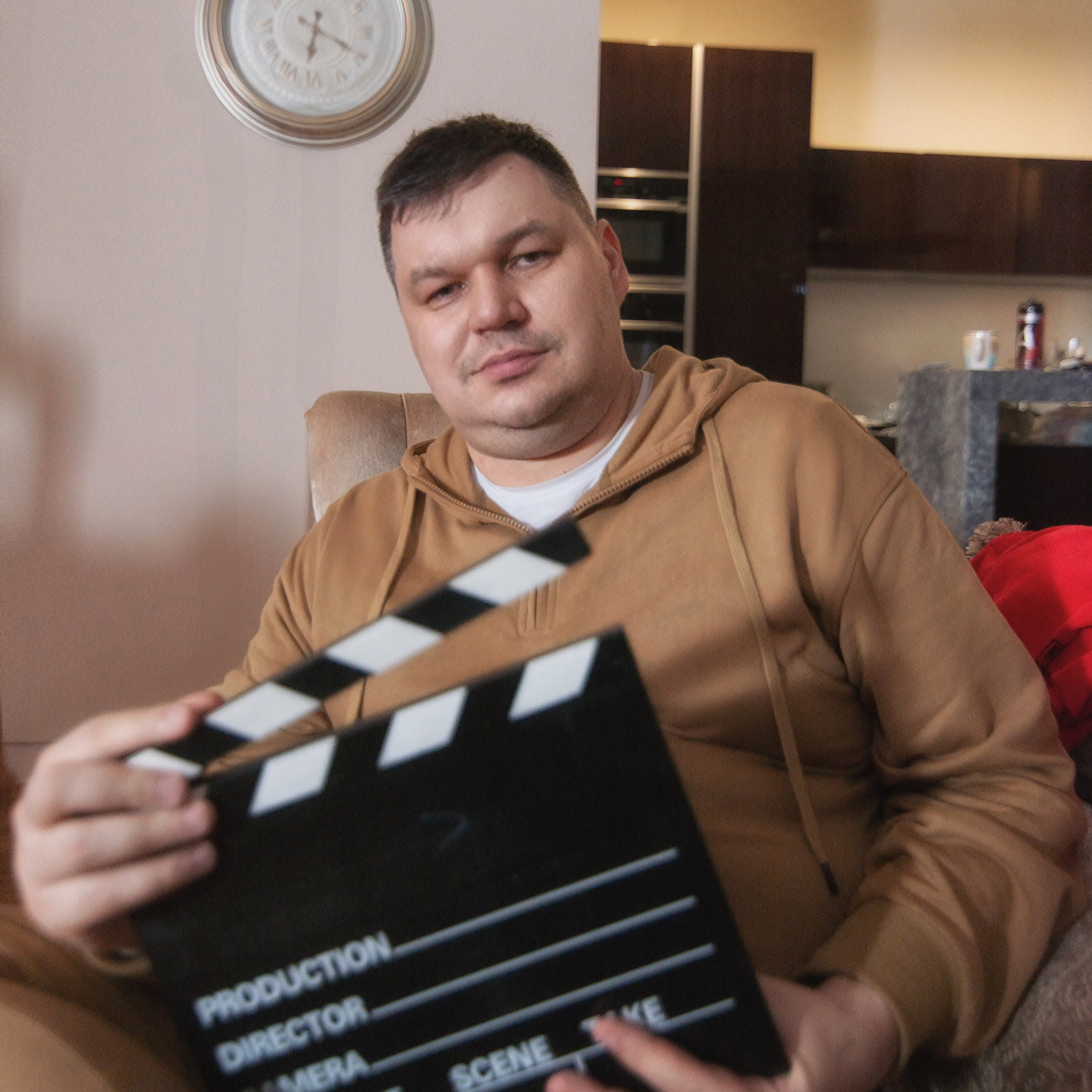 мужчина в коричневом худи, сидящий в кресле. Он держит в руках хлопушку, что создаёт атмосферу съёмочной площадки. На заднем плане видны настенные часы и кухня с тёмной мебелью. Освещение мягкое, создающее уютную обстановку.Заголовок: «Режиссёрская пауза: вдохновение для творчества»Ключевые слова: портретный фотограф, Москва, фотограф Сафронов Иван, хлопушка, кино, творчество, интерьерная съёмка.